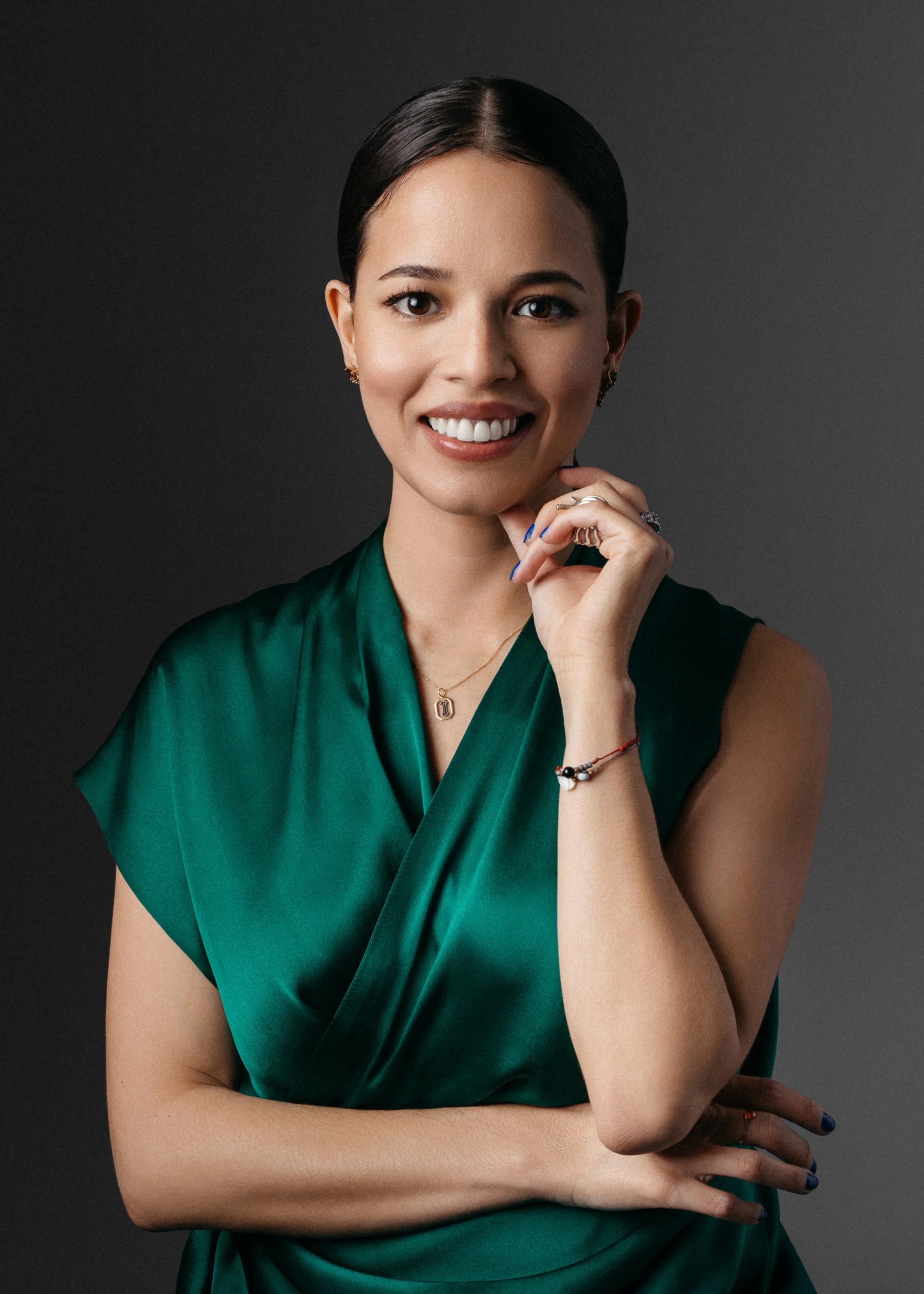 A professional corporate headshot of a woman with dark hair and brown eyes smiling, wearing a green satin dress, jewelry, and a dark background.