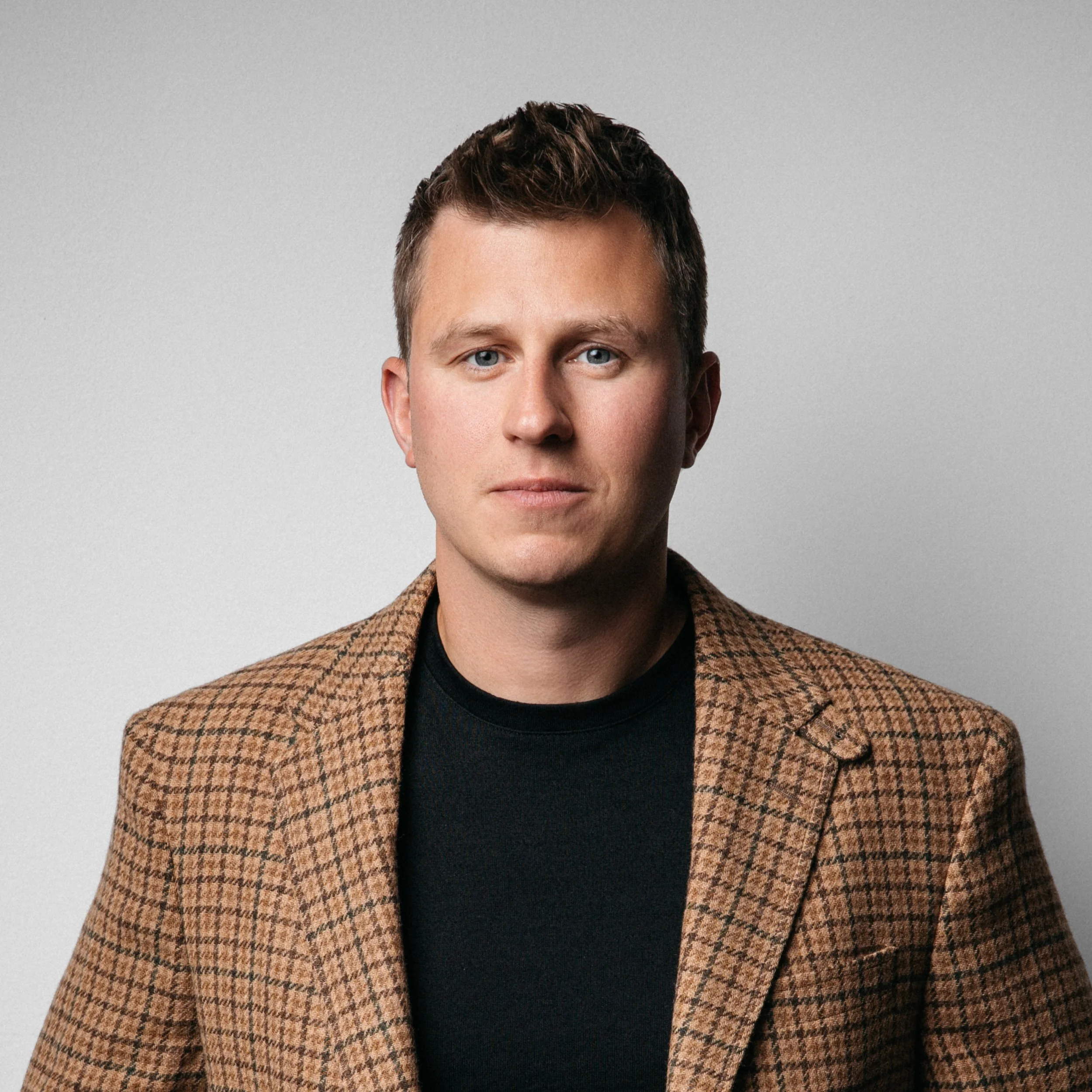A professional corporate headshot: a close-up of a person's face showing blue eyes, short brown hair, a brown checkered blazer, and a black shirt.