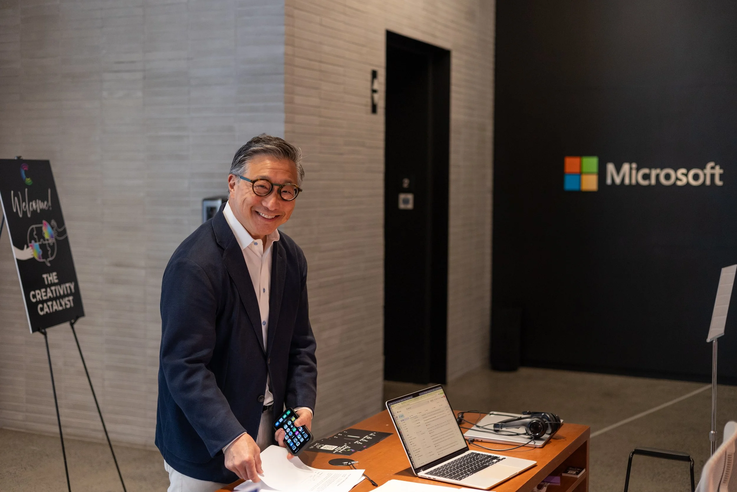 A corporate event professional photo of a smiling man in a suit holding a smartphone at a tech booth with a laptop, headphones, and marketing materials on a table. A Microsoft logo is visible on the wall behind him.