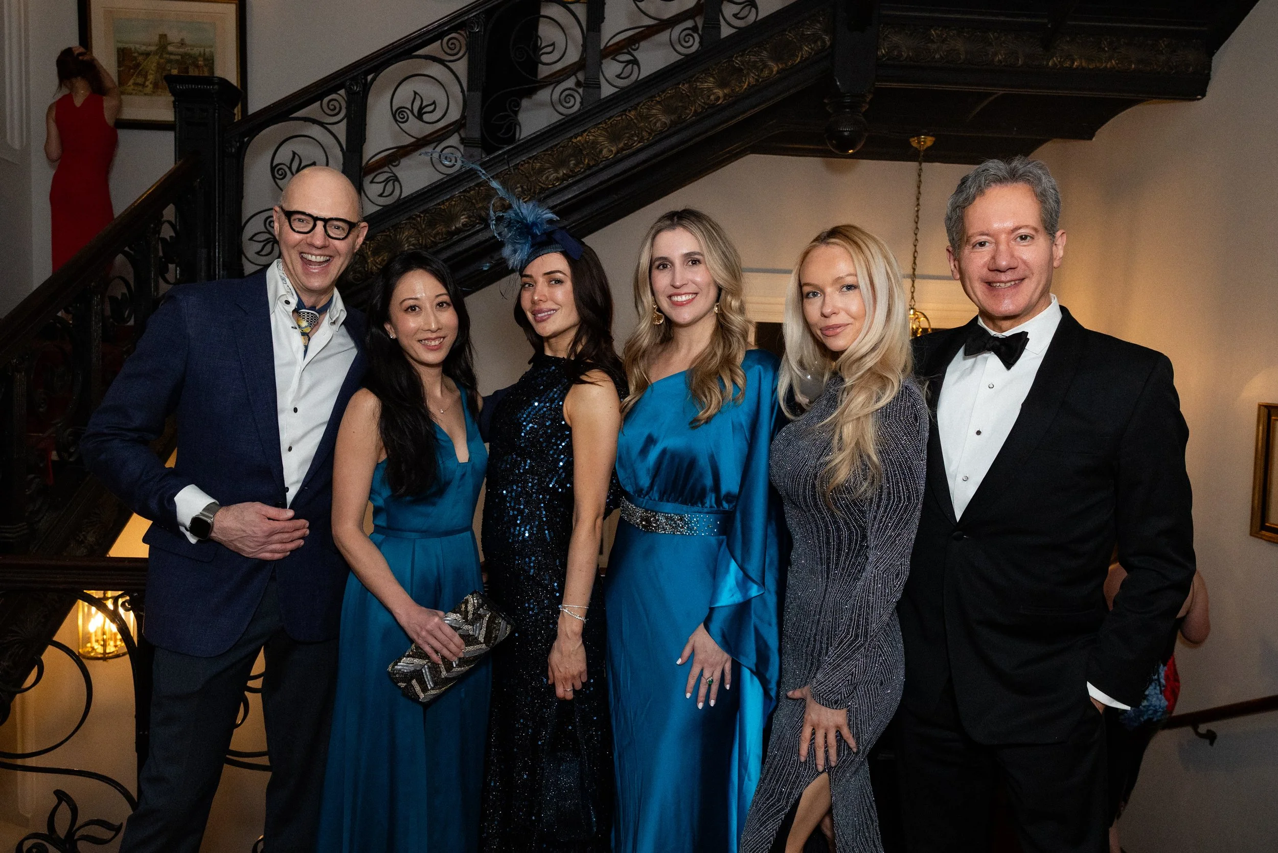 A corporate event: a group of six people, four women and two men, dressed in formal and semi-formal attire, standing together indoors near a staircase, smiling at the camera. A woman in a red dress is seen in the background near the staircase.