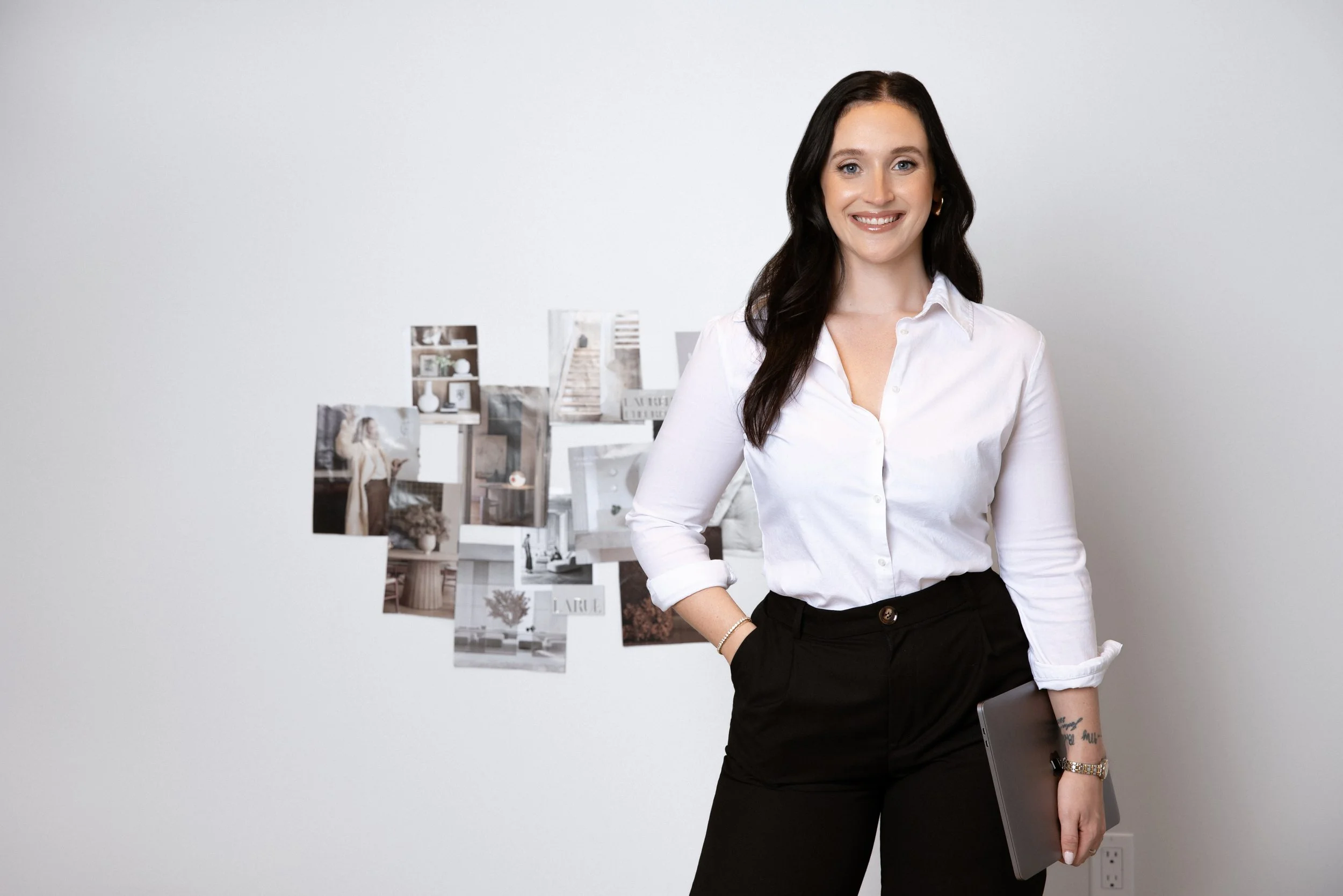 A professional corporate headshot of a woman with dark brown hair wearing a white blouse and black pants, standing in front of a white wall with a bulletin board of photos behind her, smiling, holding a gray laptop in her right hand.