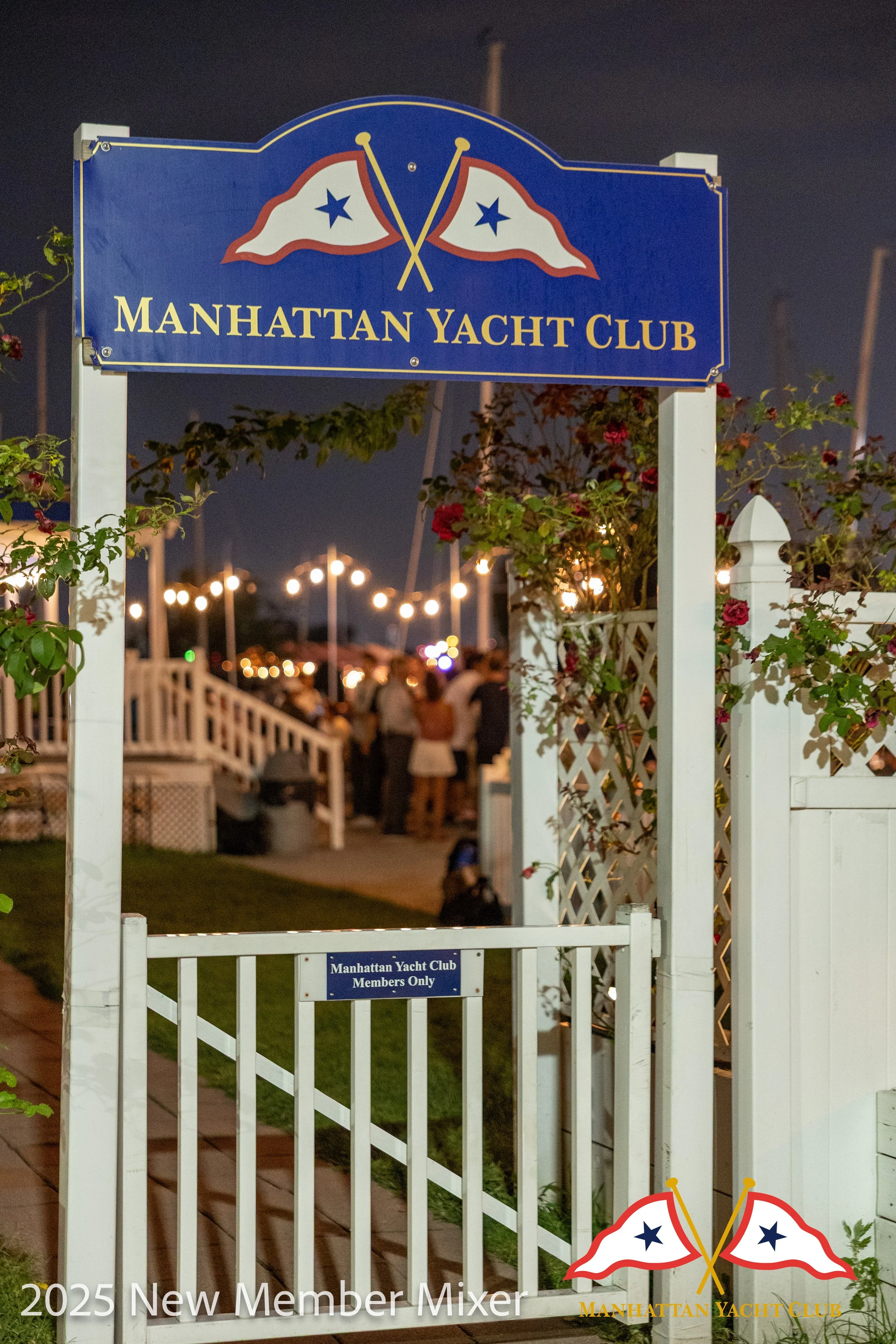A corporate event professional photo of a nighttime scene at the Manhattan Yacht Club with a white gate, a blue sign with gold text and two flags, and a crowd of people in the background. The sign reads 'Manhattan Yacht Club' and 'Members Only'.