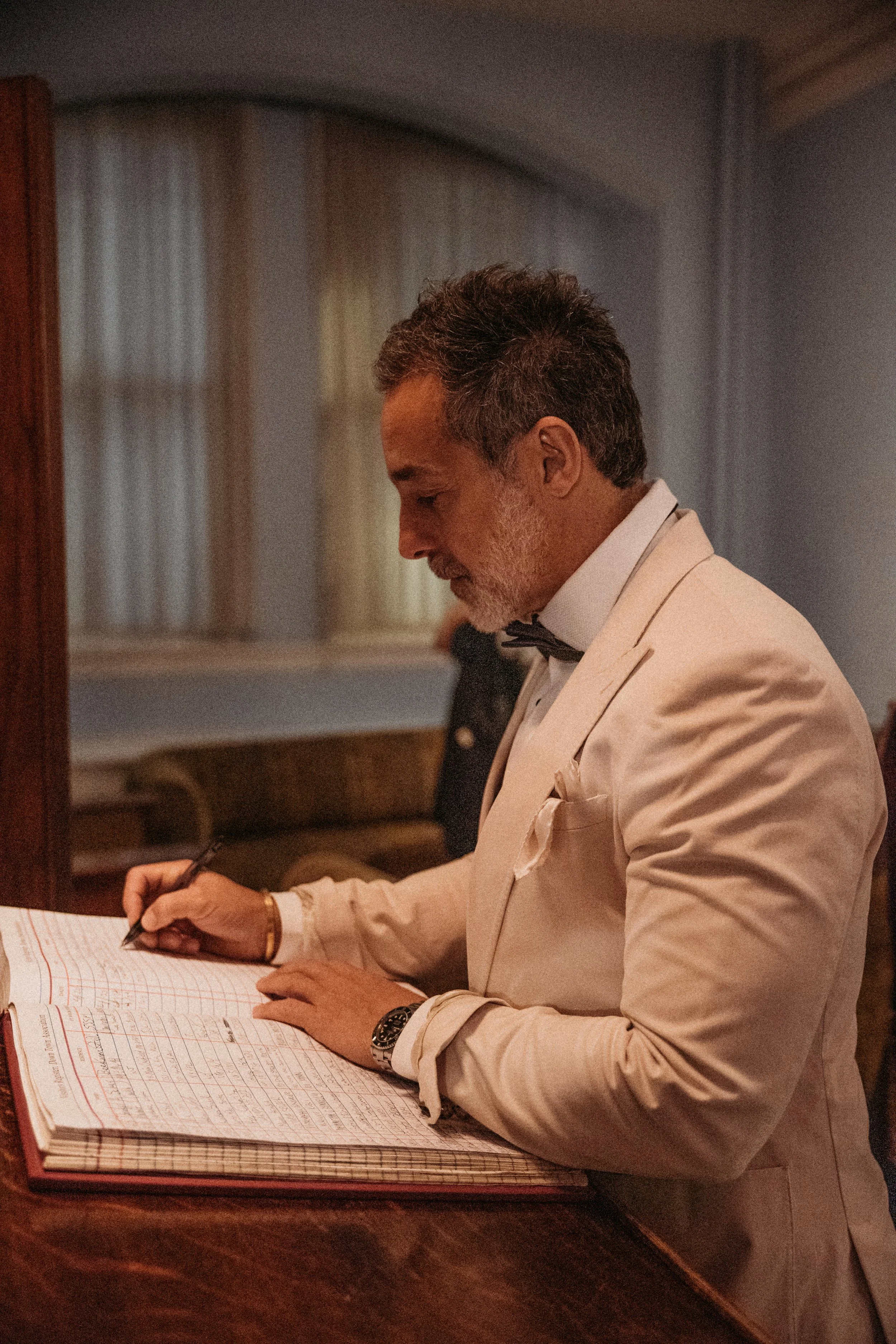A corporate event: a man in formal attire, including a white tuxedo jacket, a black bow tie, and a watch, writing in a large lined ledger or register with a pen. The room has a soft, warm ambiance with muted colors and curtains in the background.