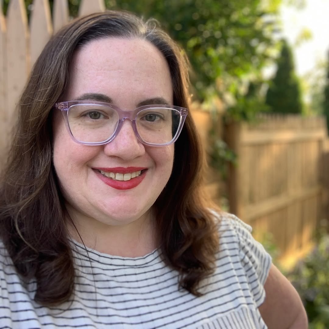 A woman with brown hair, wearing glasses and a striped shirt, smiling outdoors near a wooden fence with greenery.