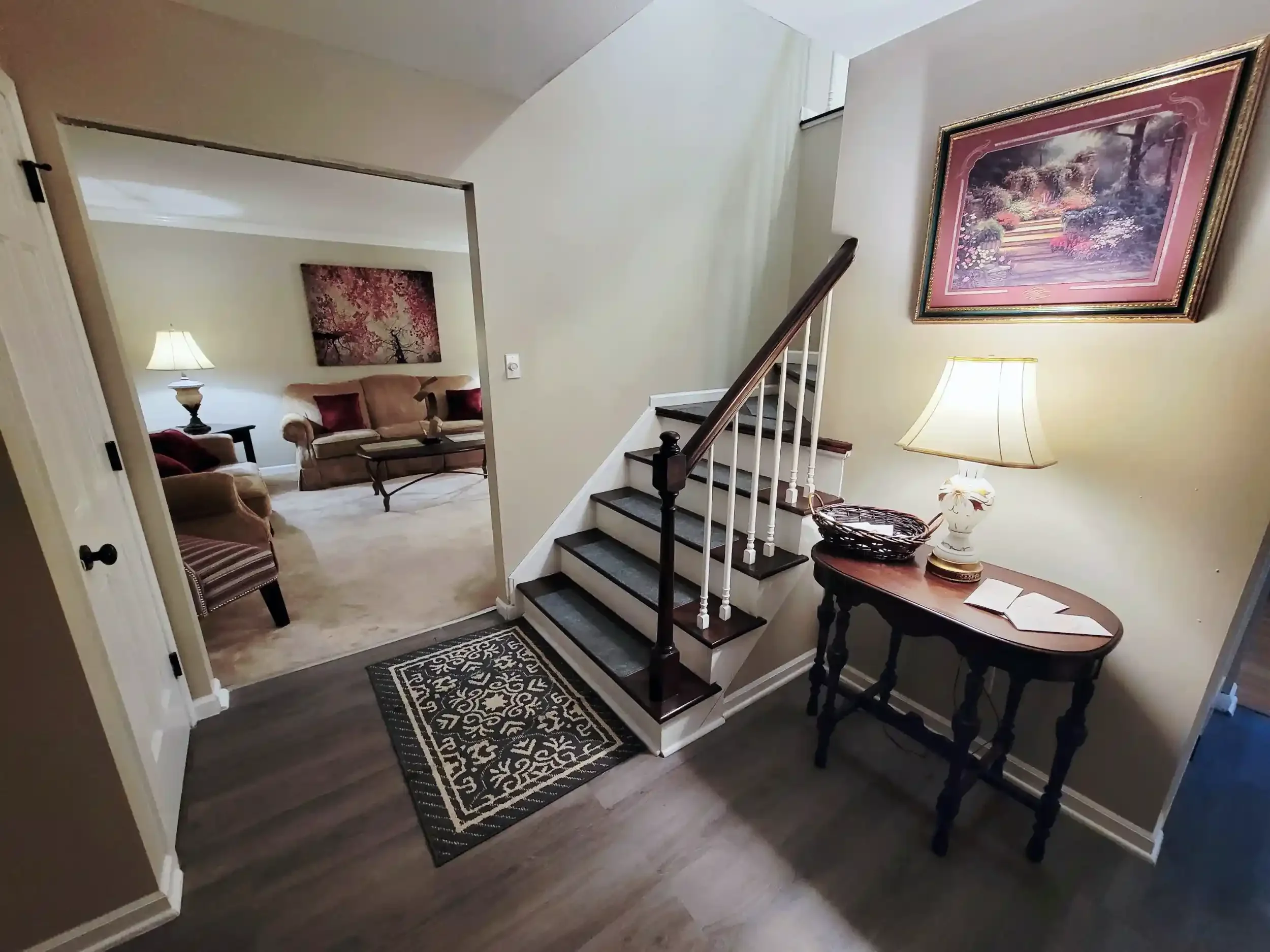 Interior view of a home with a staircase, a table lamp, and framed artwork on the walls, leading into a sitting area with sofas and a painting of pink trees.
