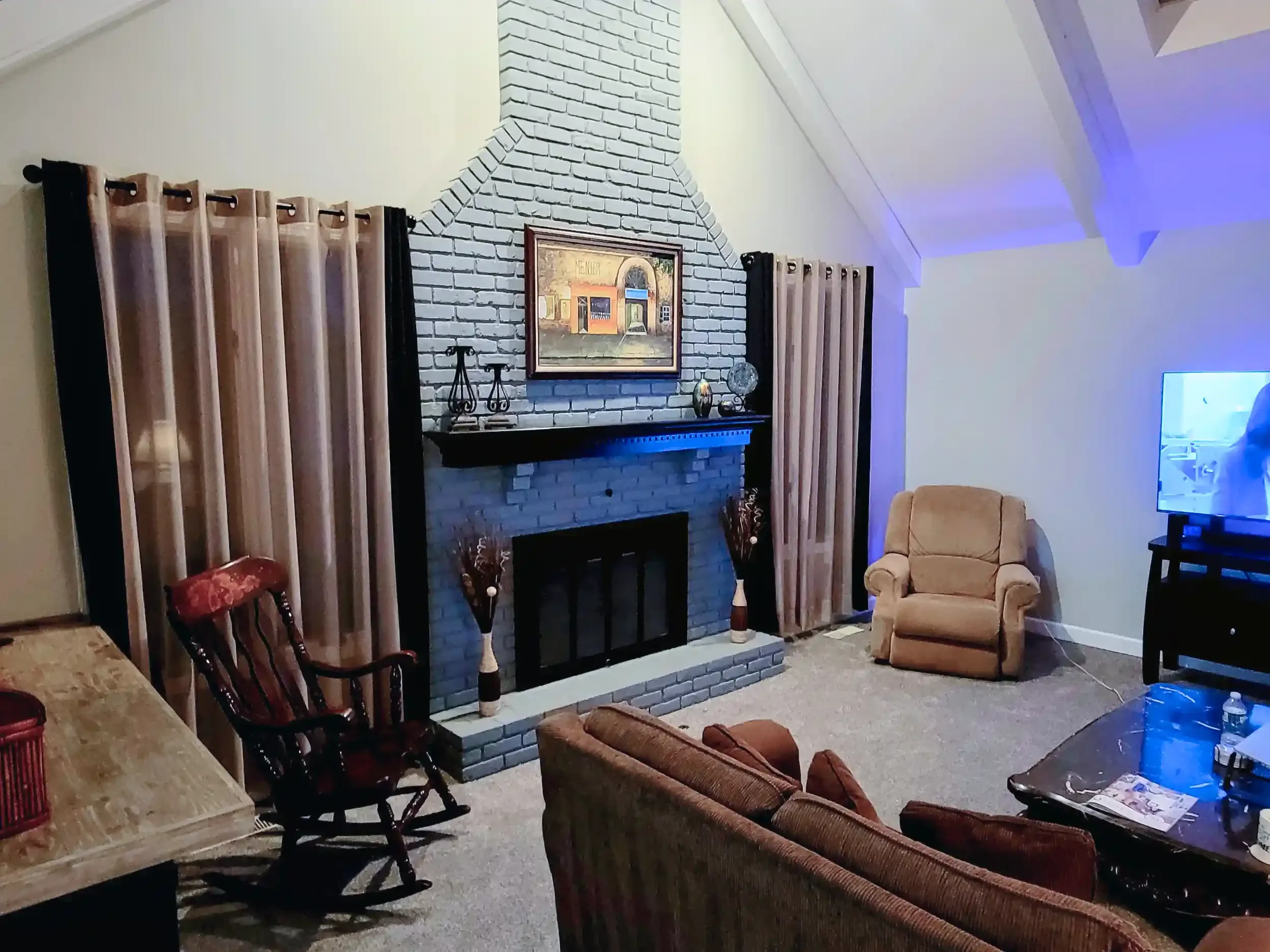 Living room with a brick fireplace, framed painting above the mantel, beige armchair, and a wooden dining table with chairs. Curtains are on both sides of the fireplace, and a television is in the corner.