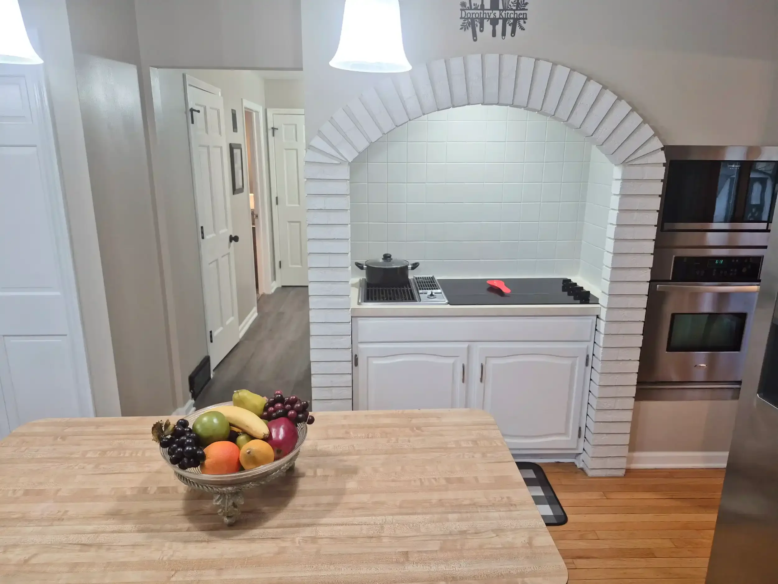 A kitchen with white cabinetry and a brick archway. A wooden dining table with a bowl of assorted fresh fruits is in the foreground. There is a white tile wall with a pot on the stove, and a sign on the wall that says 'Dorothy's Kitchen'.
