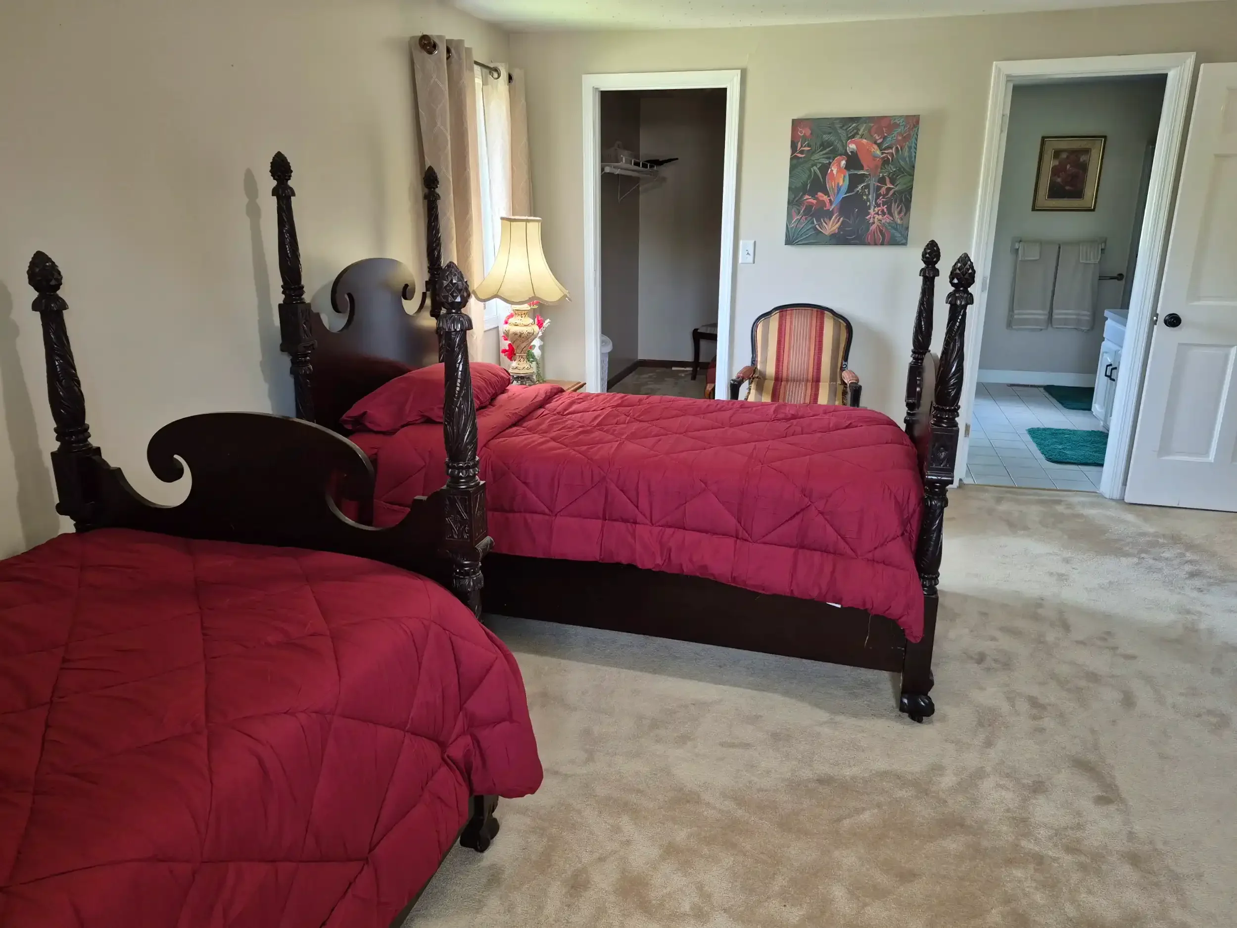 Room with two dark wooden beds with carved posts, red quilted bedding, a side table with a floral lamp, a striped chair, a painting of birds, and open doors leading to a closet and bathroom.