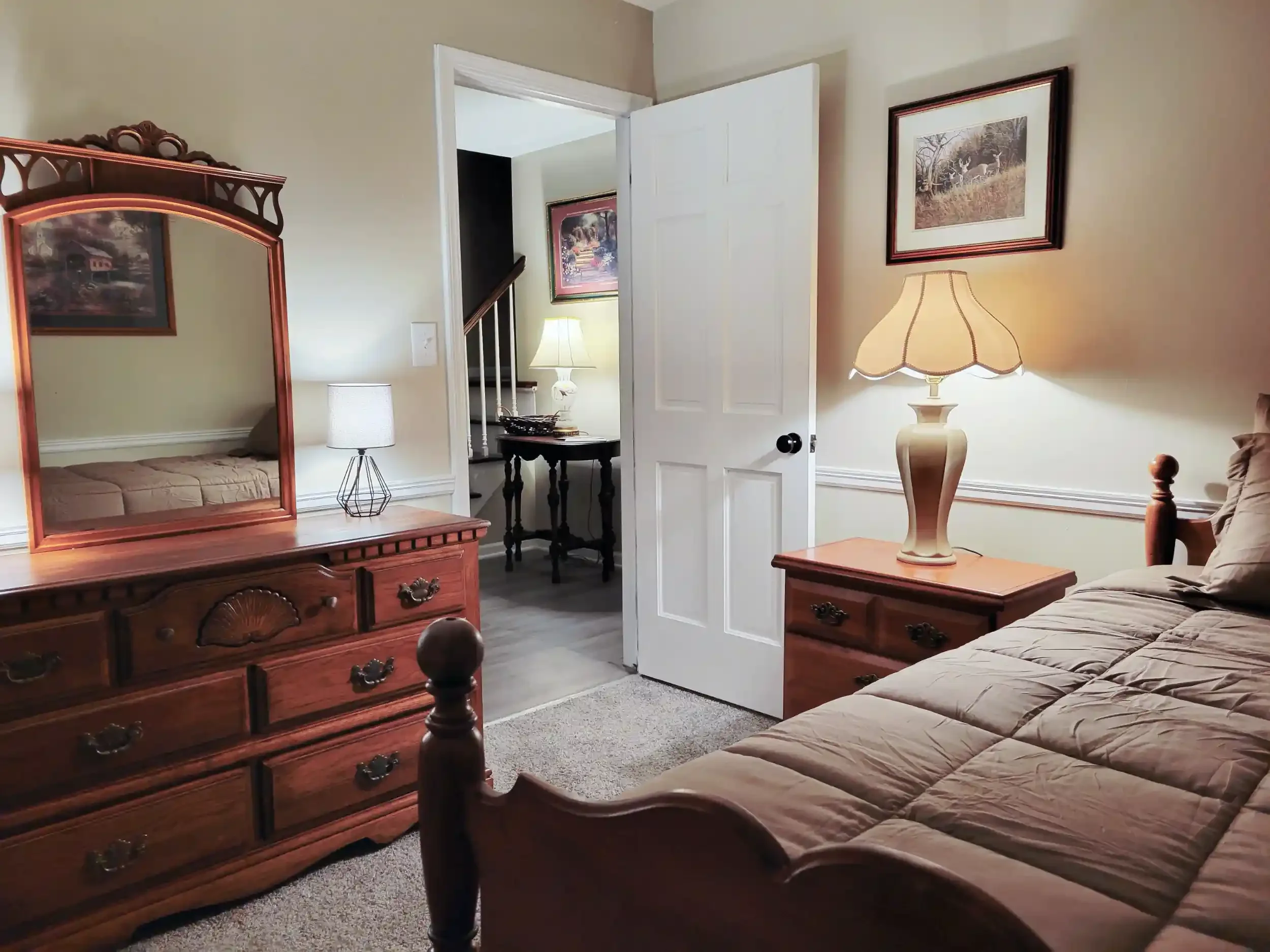 A bedroom with a wooden dresser with a mirror, a bed with a quilt, a nightstand with a lamp, pictures on the wall, and an open door leading to a hallway with a table and lamp.