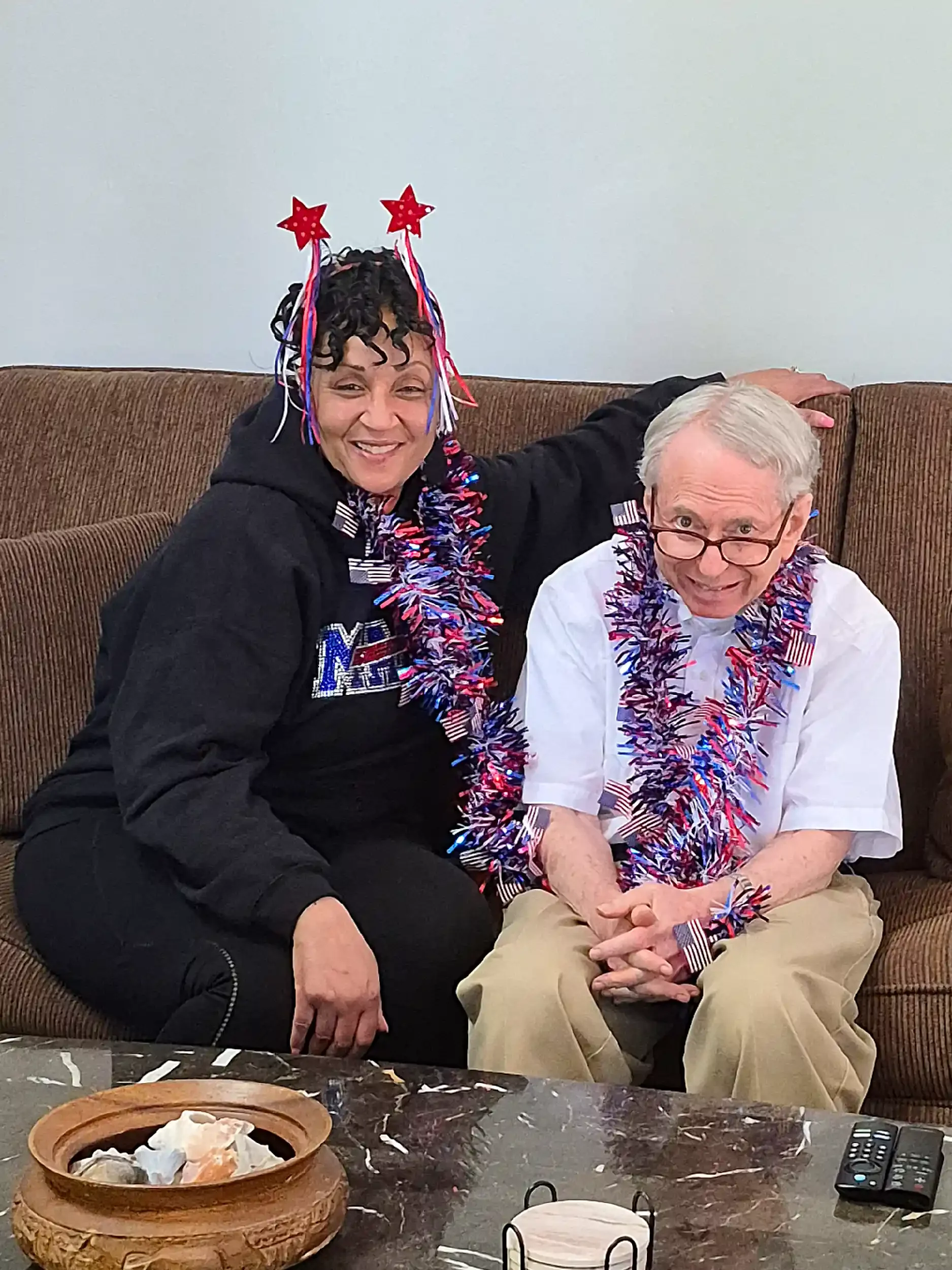 An elderly man and a middle-aged woman sitting on a brown couch decorated with patriotic and festive accessories, celebrating a patriotic holiday. The woman is wearing star-shaped decorations on her head and American flag-themed tinsel, while the man is wearing similar decorations and glasses. There is a bowl of dessert on the table in front of them and remote controls on the table.