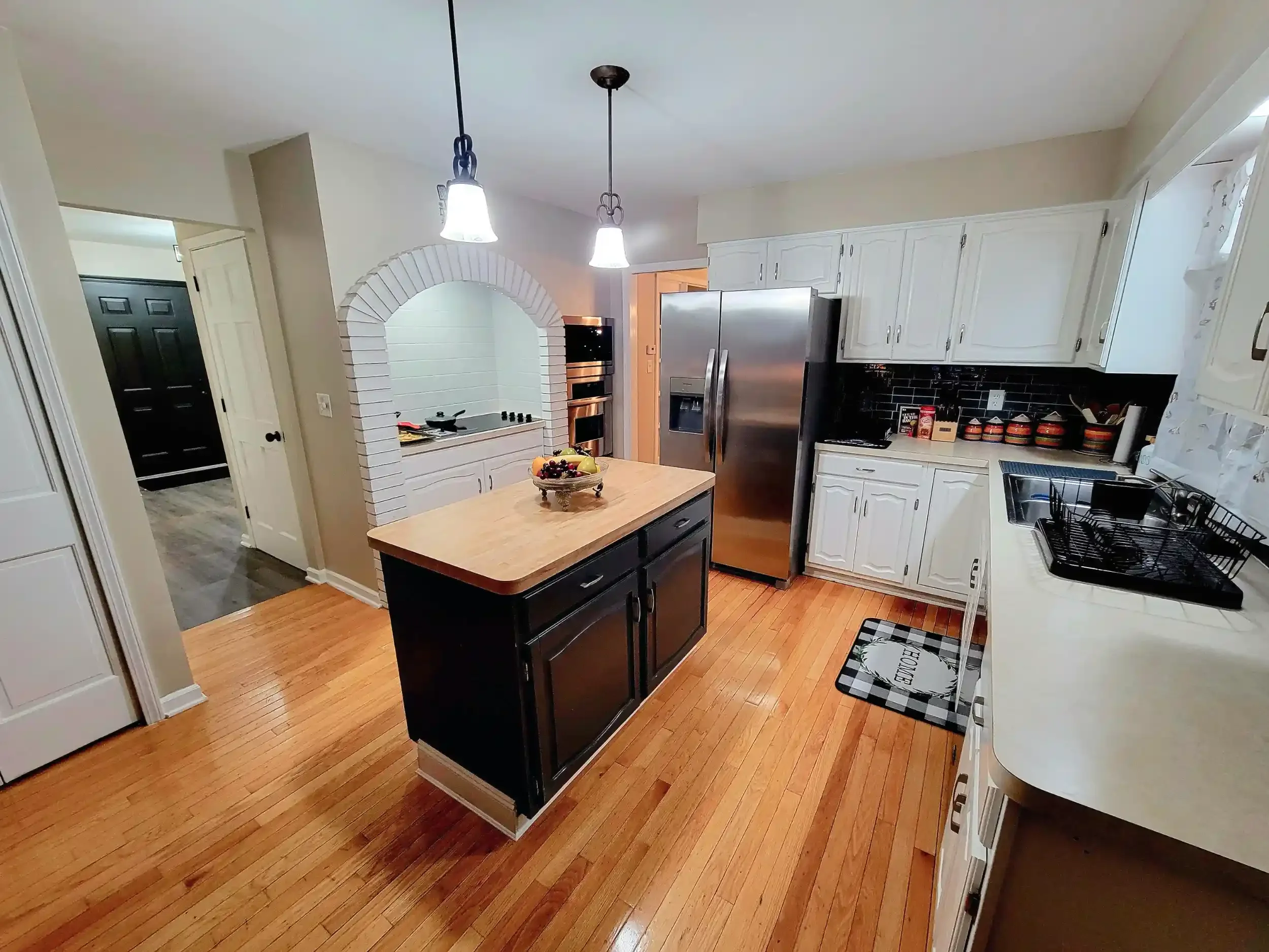 Kitchen with white cabinets, stainless steel refrigerator, black and white checkered rug, wooden flooring, black kitchen island, fruit bowl on island, black dish drying rack, and pendant lights hanging from the ceiling.