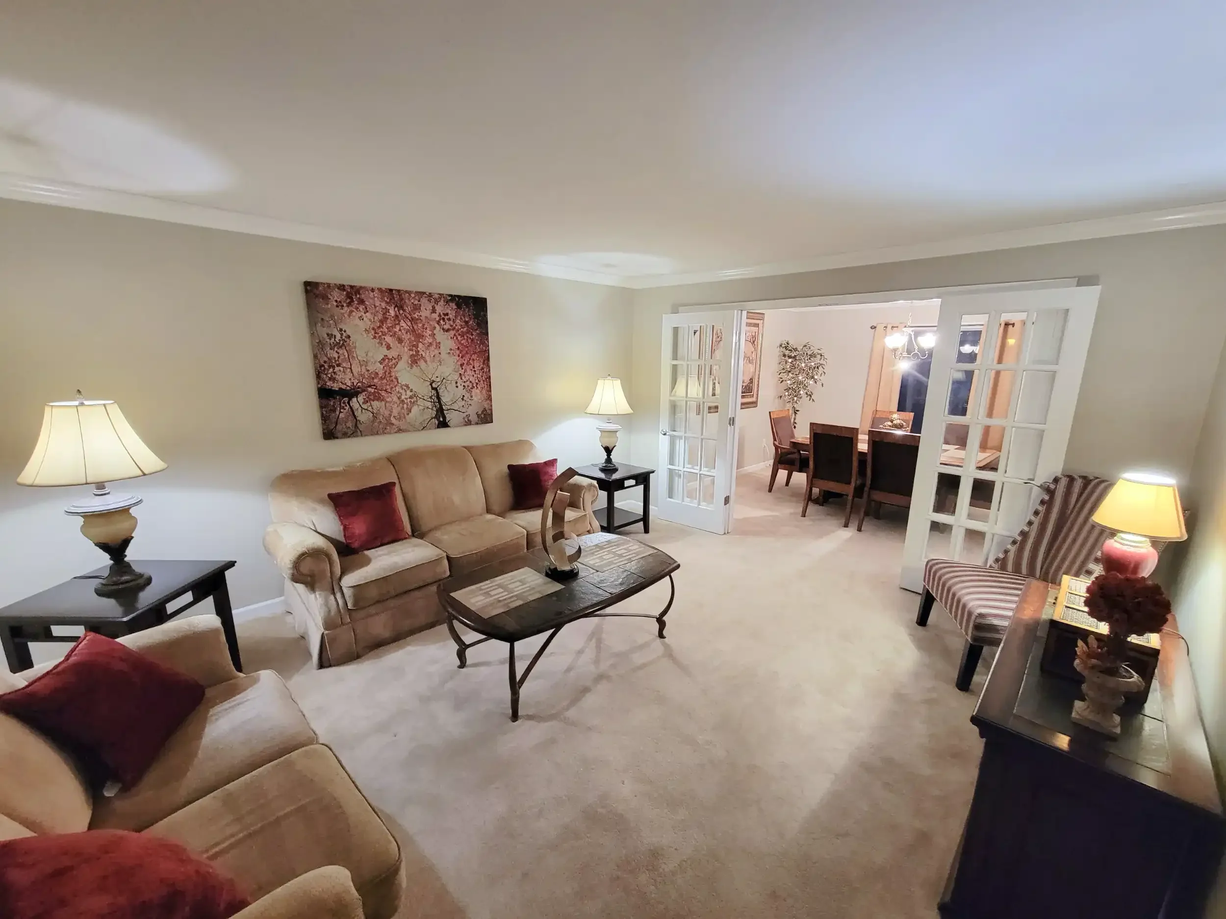 Living room with beige sofa, side tables with lamps, wall art, and a view into the dining area with table and chairs.