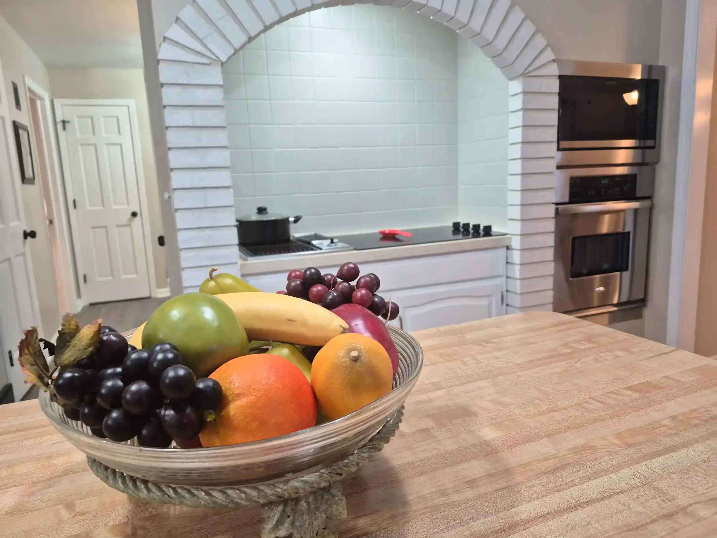 A glass bowl filled with assorted fruits including grapes, apples, oranges, and a lemon, placed on a wooden table in a kitchen with white cabinetry and appliances.
