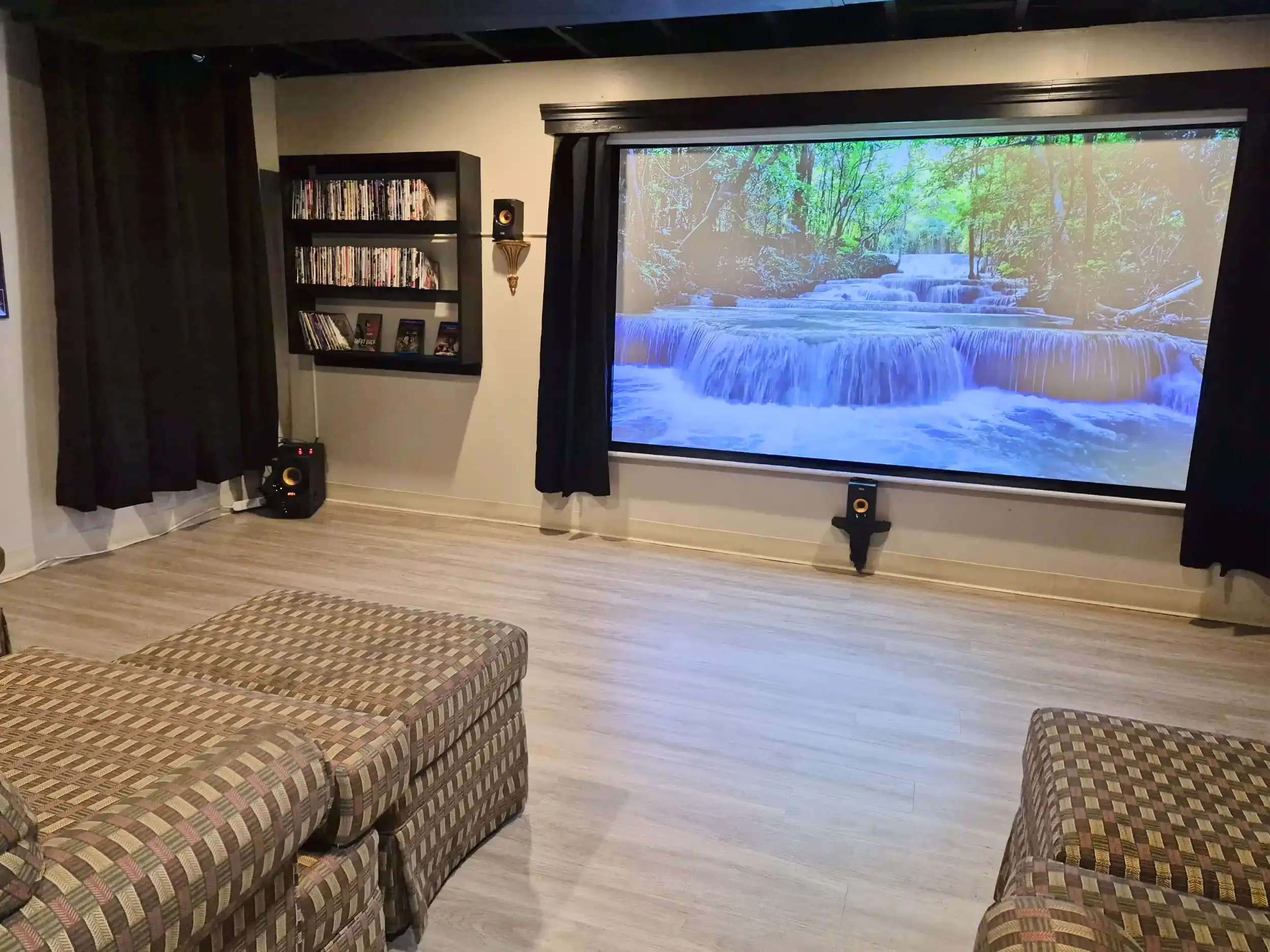 Indoor room with a wall-mounted bookshelf filled with DVDs, a wall sconce, and a large projector screen displaying a nature scene with a waterfall and green trees. There are two plaid sitting chairs and a wooden floor.