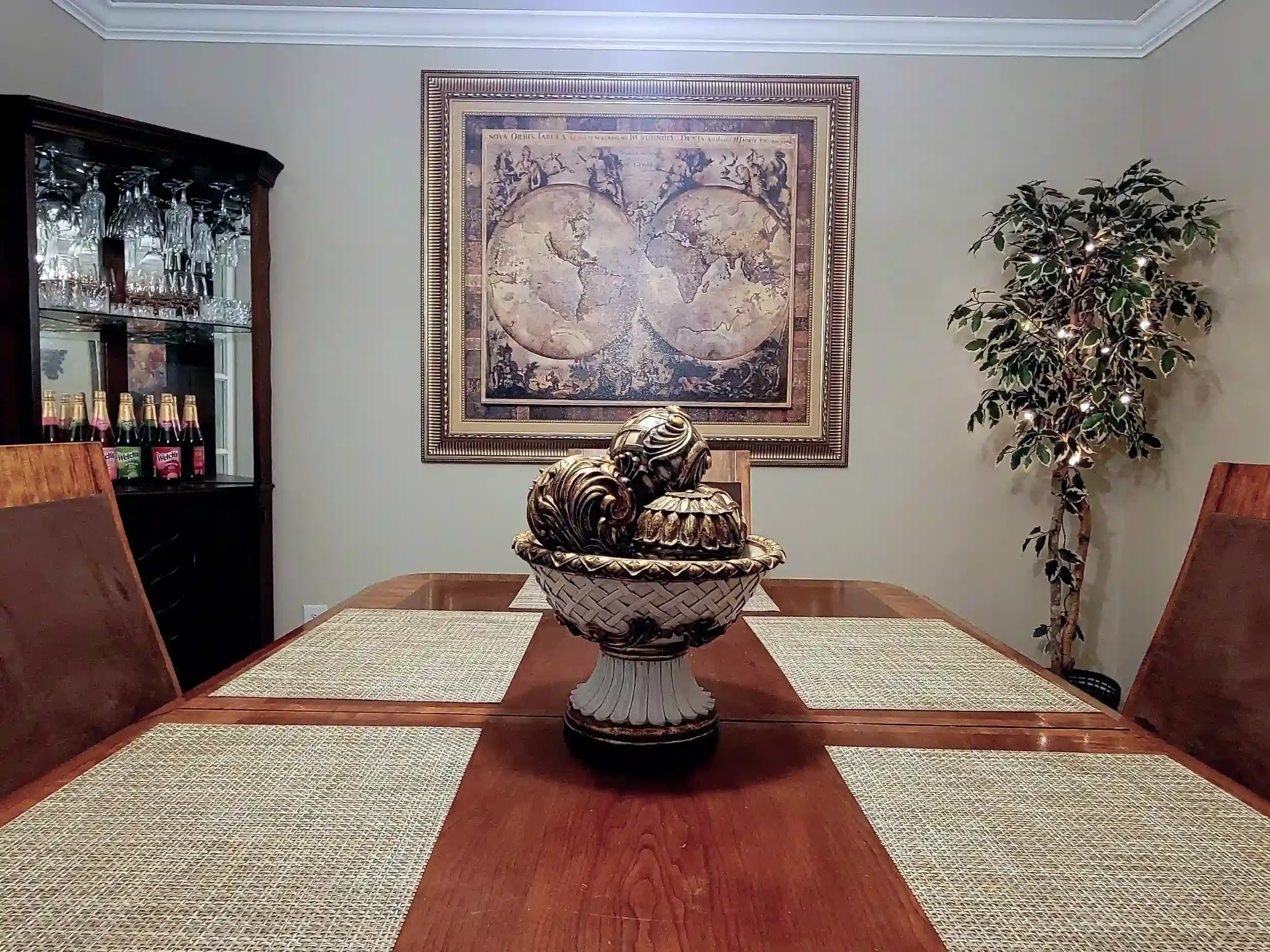 A dining room with a wooden table, featuring a decorative bowl centerpiece. The table has woven placemats and wooden chairs. On the left, there is a glassware cabinet with wine glasses and bottles. The wall is decorated with a large framed world map. To the right, there are potted plants with lights, adding a touch of greenery to the room.