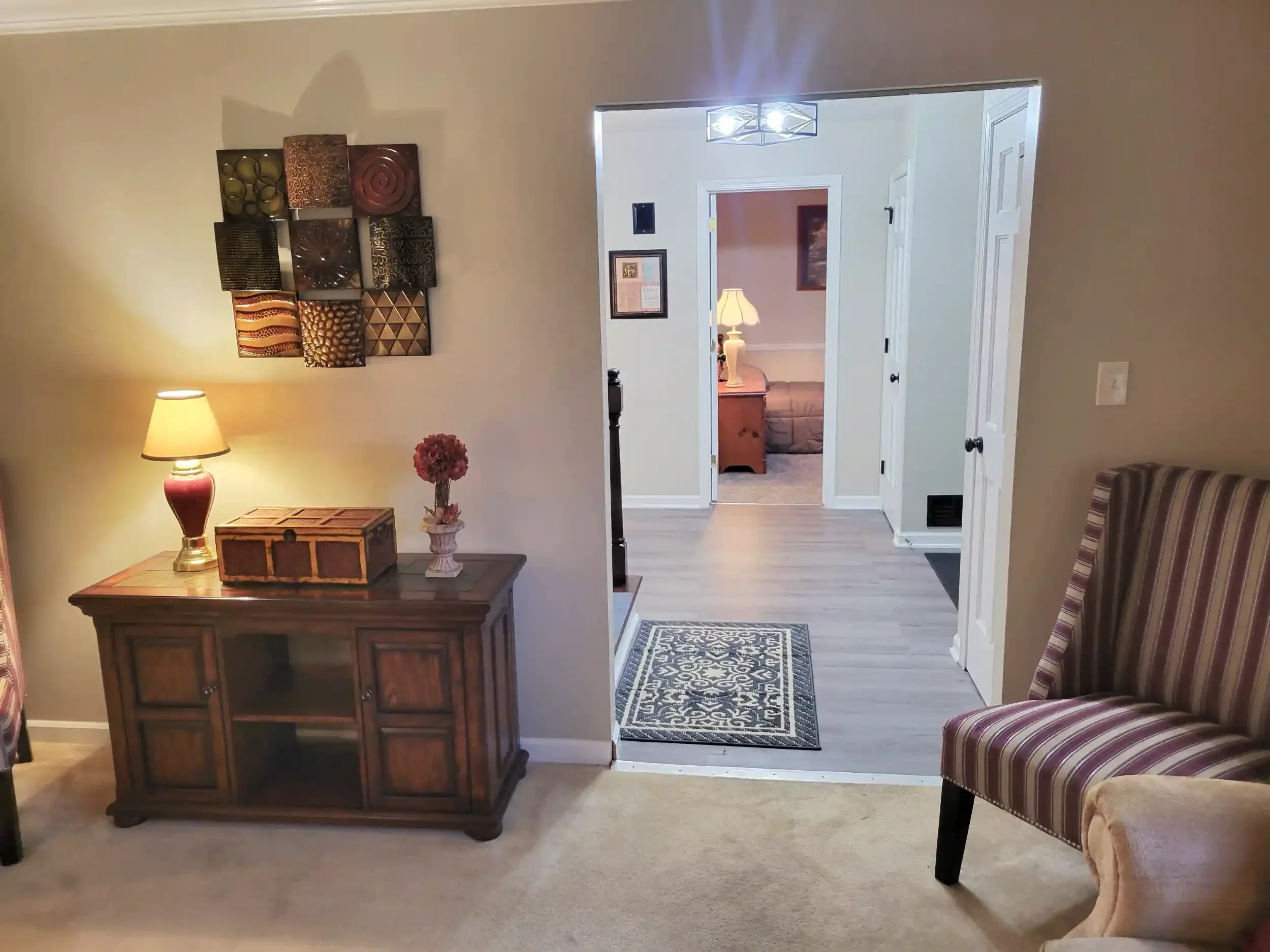 View of a living room with a wooden sideboard, a lamp, a vase of pink flowers, a patterned area rug, and a striped armchair. A decorative wall hanging and a doorway leading to a room with a lamp and sofa are visible.