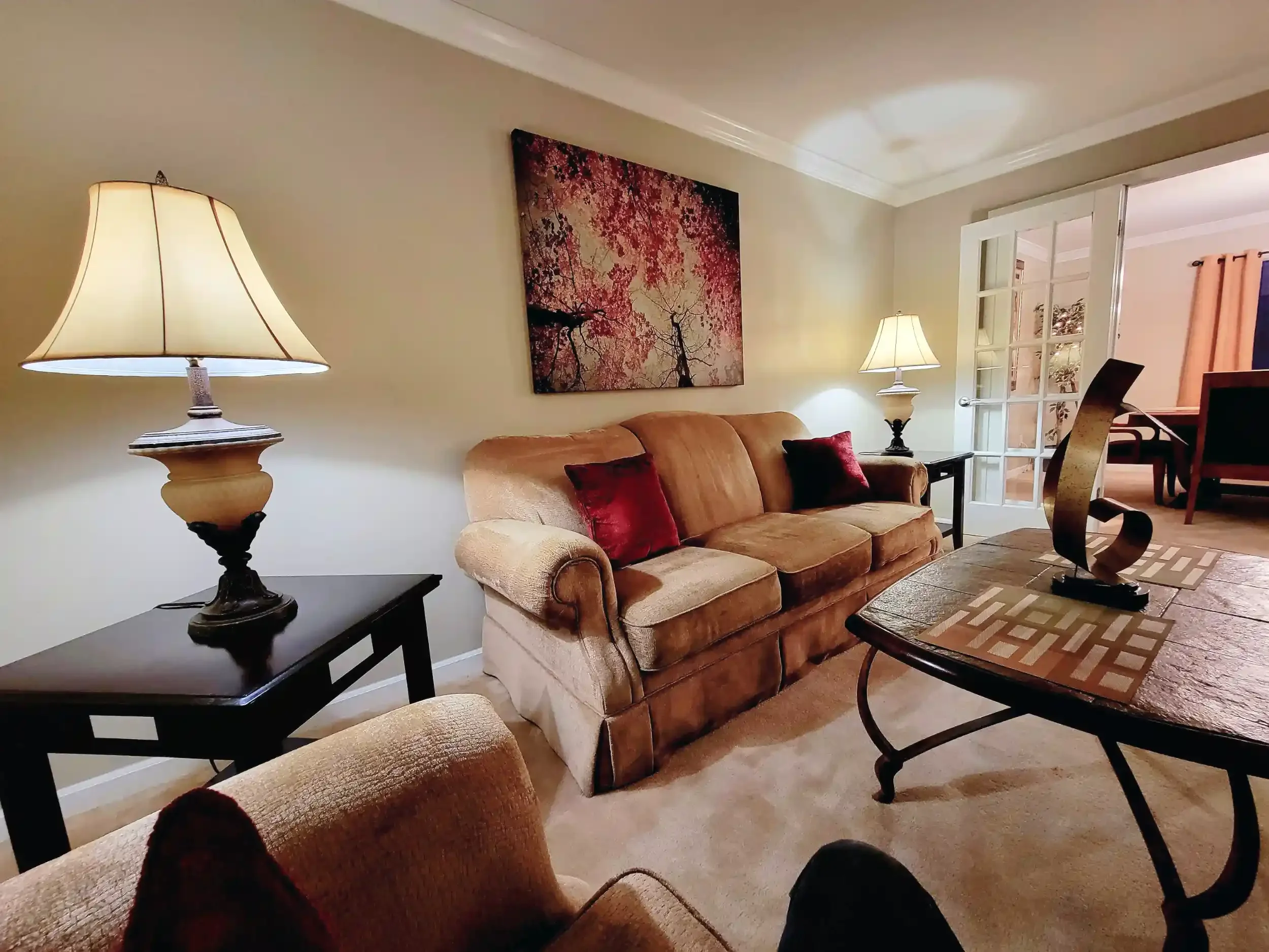 Living room with beige sofa adorned with red cushions, antique-style black side tables, two lamps with beige shades, a wall painting of pink cherry blossoms, a wooden dining table with chairs, and a glass-paneled door opening to another room.