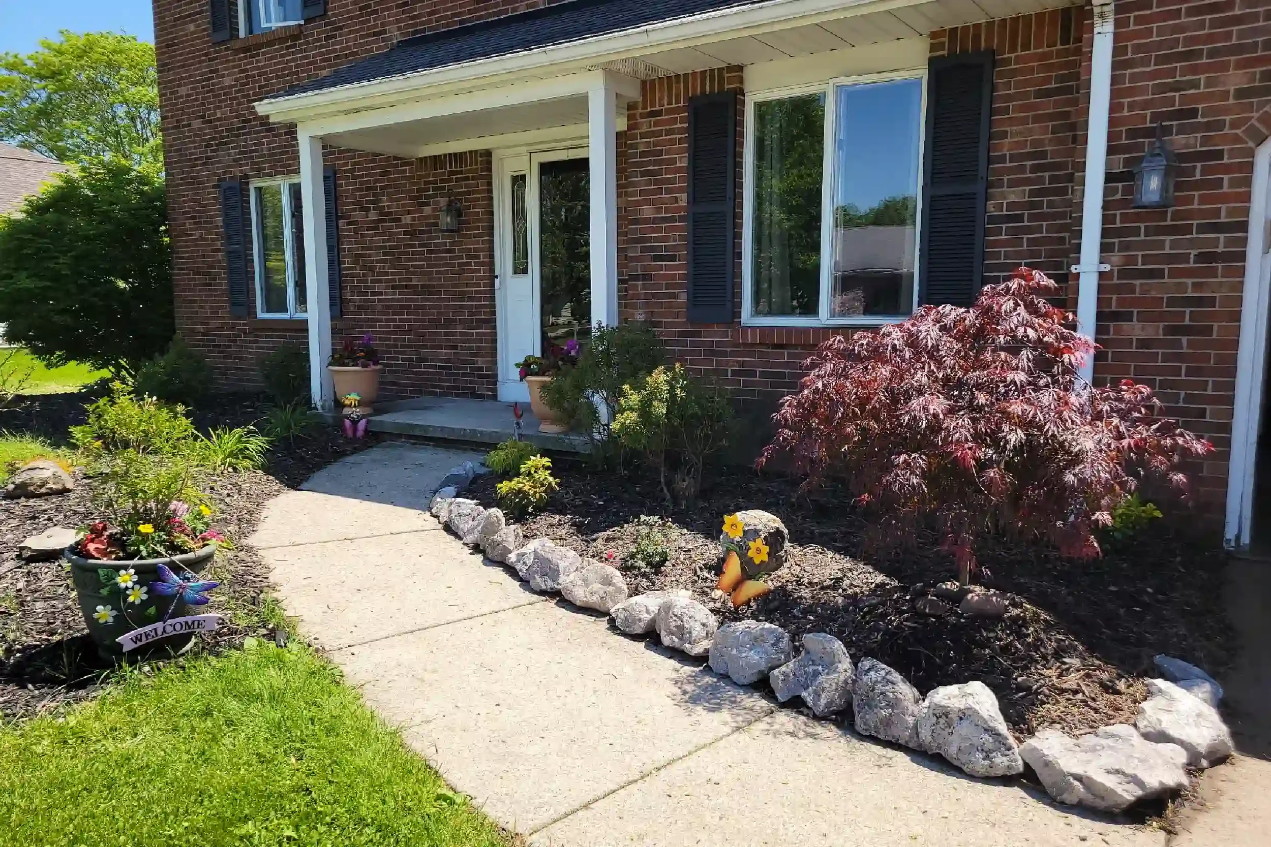 Front entrance of a brick house with a small porch, black shutters, and a glass front door. The porch has potted plants with flowers. The front yard has a flower bed with rocks, colorful flowers, and a small shrub. A concrete walkway leads to the por