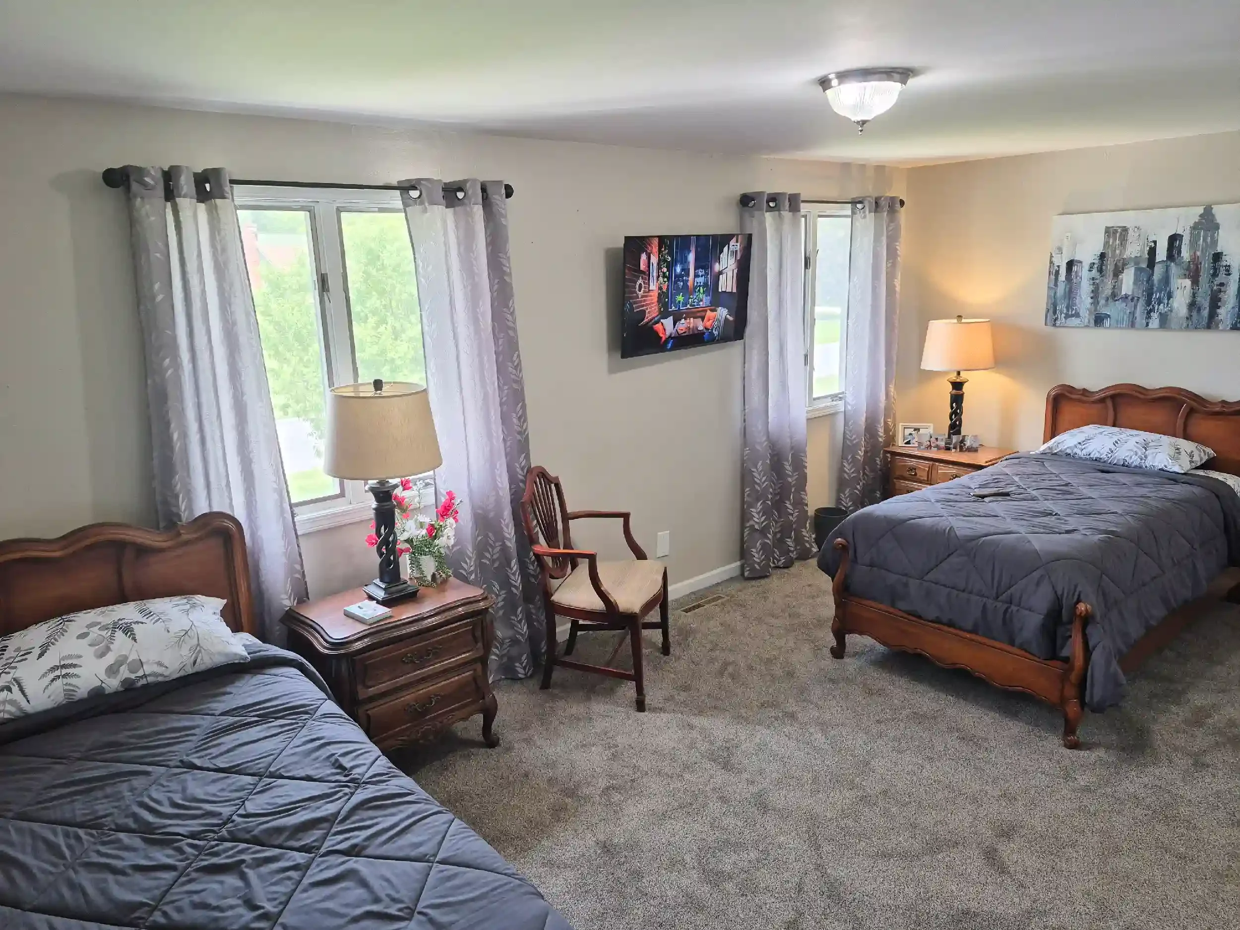 A bedroom with beige walls, two windows with white and patterned curtains, a wooden bed with a gray quilt, two wooden nightstands with lamps, a wall-mounted TV, and framed artwork.