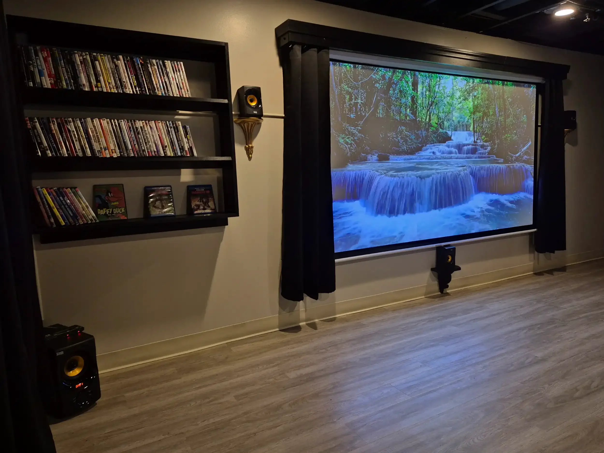 A living room with a large projection screen showing a waterfall in a forest. To the left of the screen, there are two shelves filled with DVDs and VHS tapes. There are two speakers, one next to each side of the screen, and a small subwoofer on the f