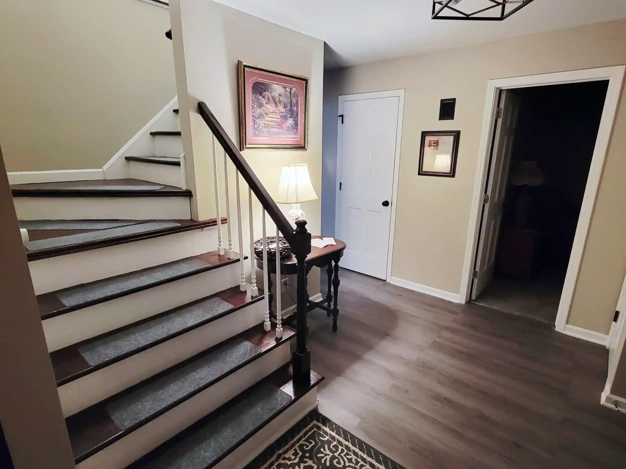 Interior view of a home with stairs, a lamp on a small table, framed pictures on the wall, and a door leading to a dark room.