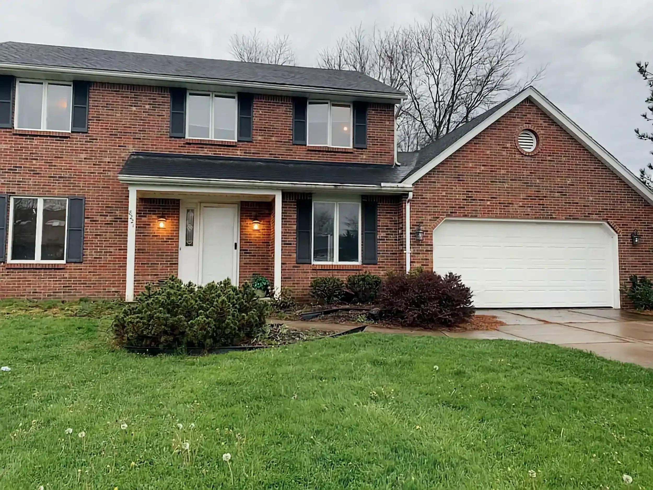 A red brick house with black shutters, a white garage door, and a front porch with two outdoor lights. The lawn is green with a few bushes in front, and there are bare trees and cloudy sky in the background.