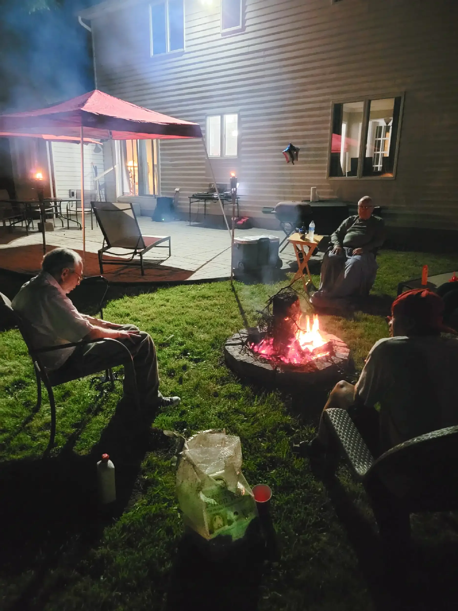 People sitting around a campfire in a backyard at night, with a house and patio visible in the background.