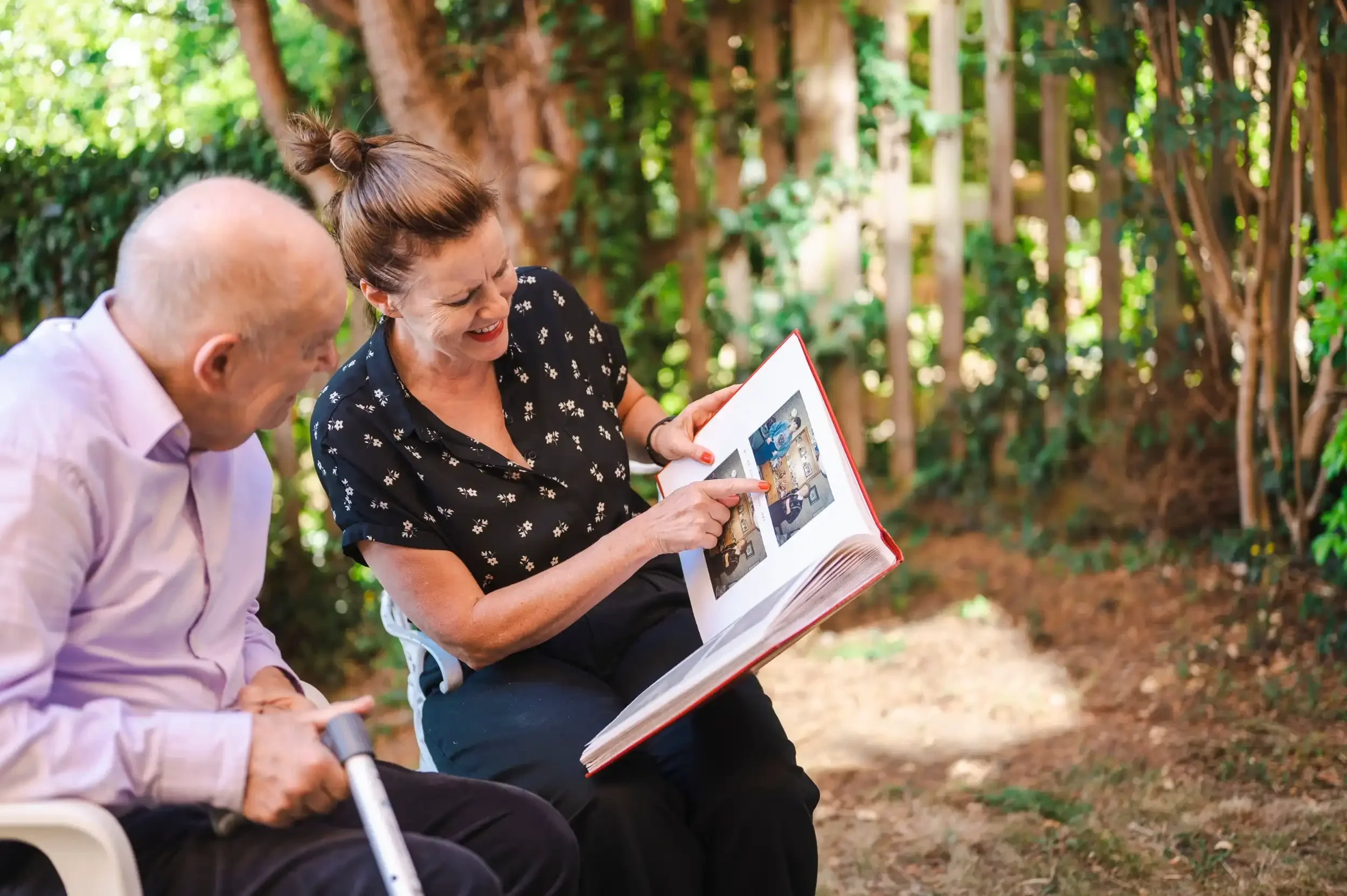 Older woman and man sitting outside in a wooded area, reading a photo album together, smiling and enjoying the moment.