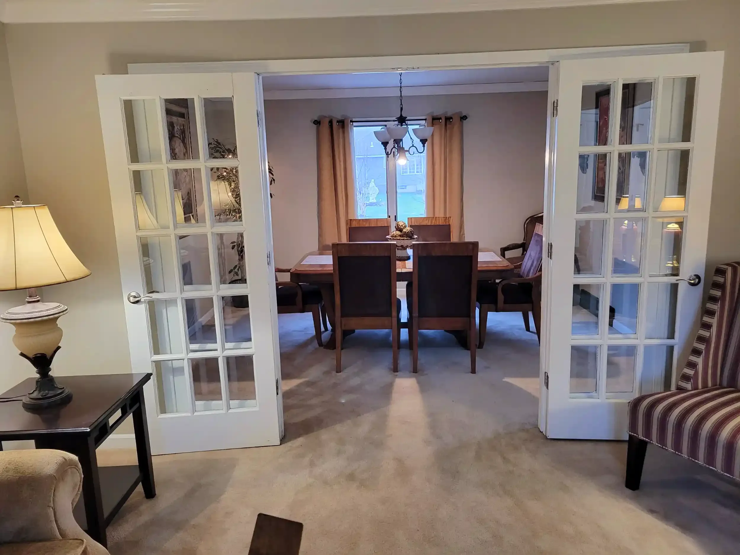View through glass-paneled double doors into a dining area with a wooden table and chairs, curtains on a window, and a chandelier hanging from the ceiling.