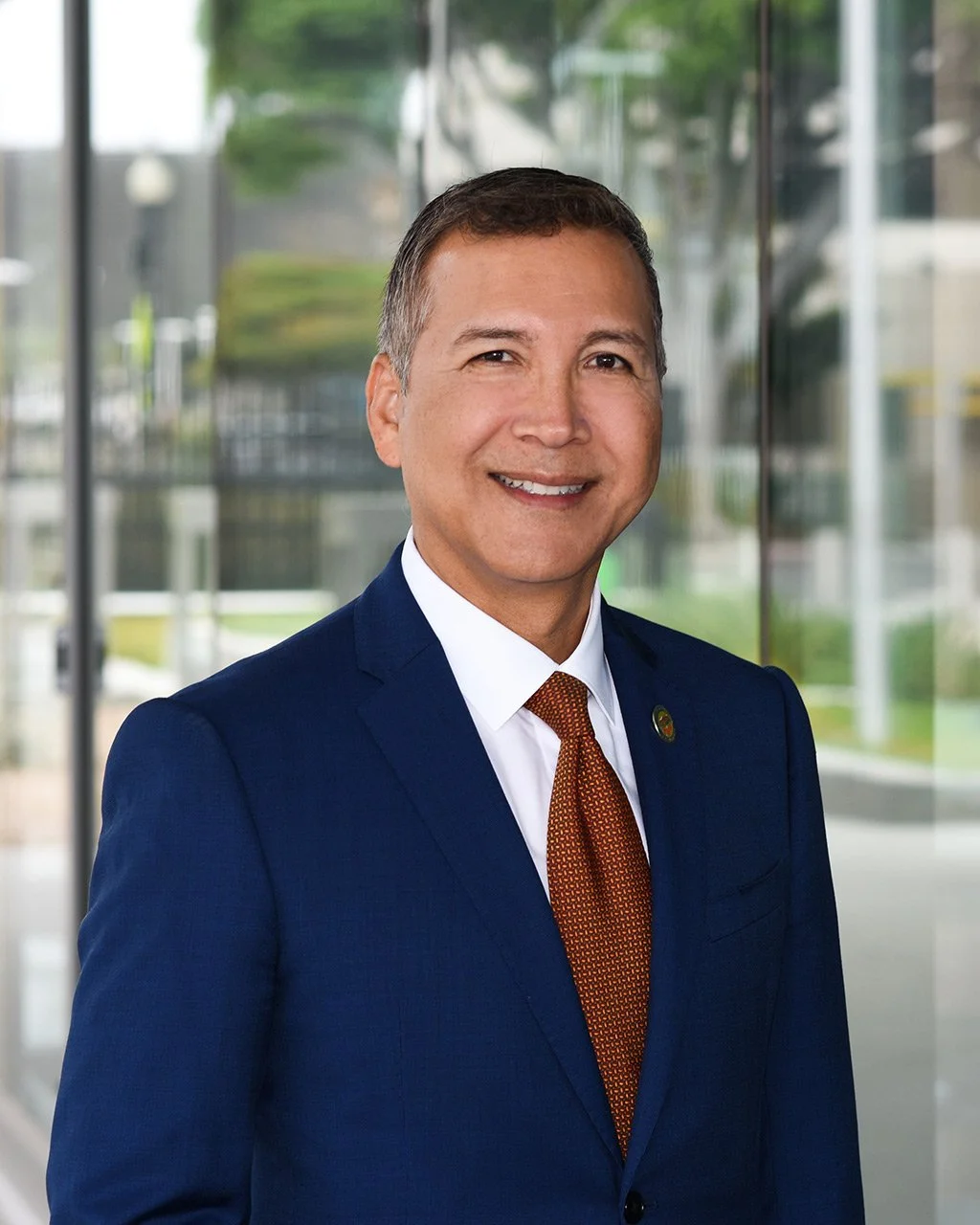 A smiling man in a navy suit with a white shirt and orange tie standing indoors with glass walls and an outdoor view.