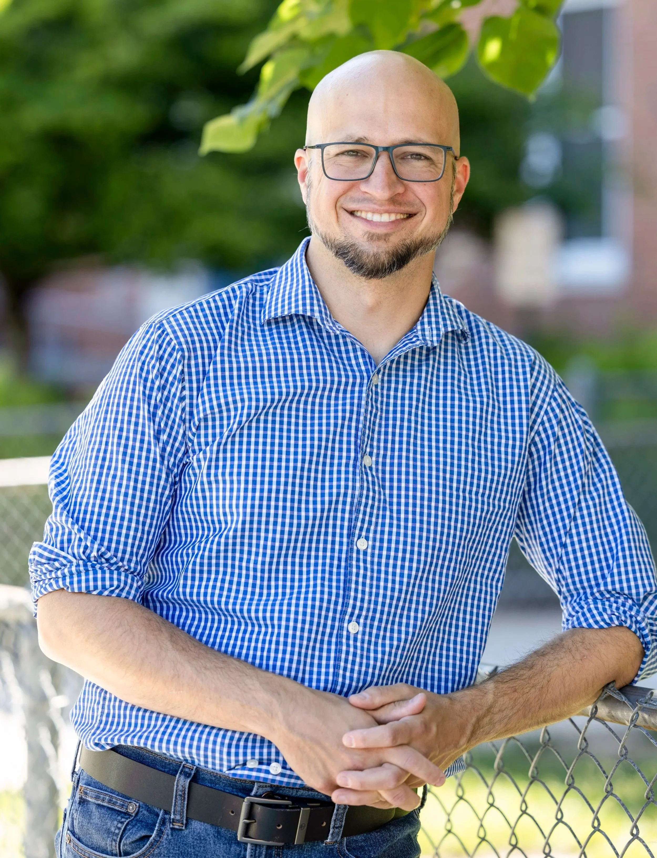 A man with glasses, a trimmed beard, and a shaved head wearing a blue checkered shirt and jeans, standing outdoors and leaning on a chain-link fence with greenery in the background.