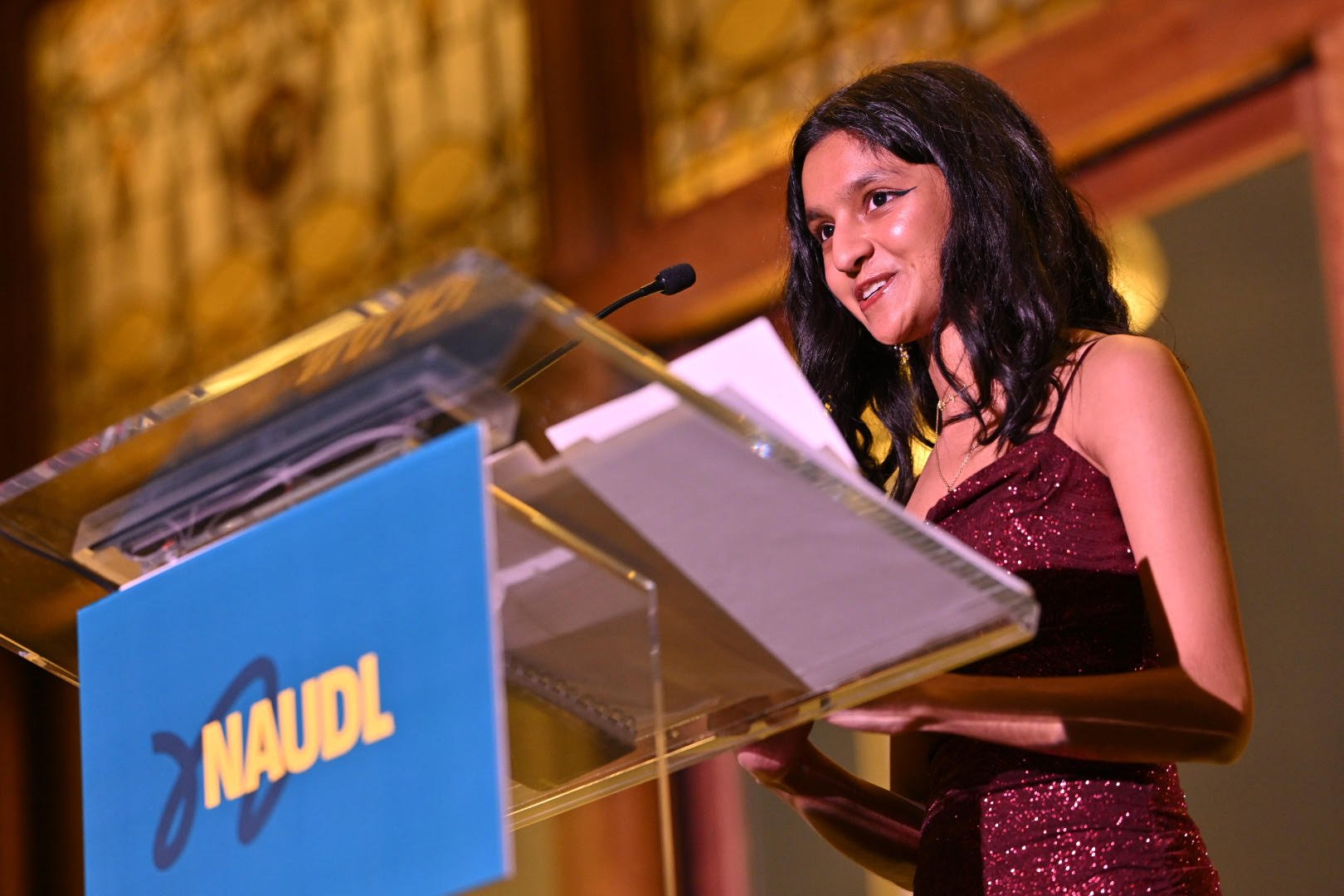 A woman in a sparkling burgundy dress speaking at a podium with a blue sign that reads "NAUDL." She has shoulder-length black hair and is smiling, standing in a warmly lit room.