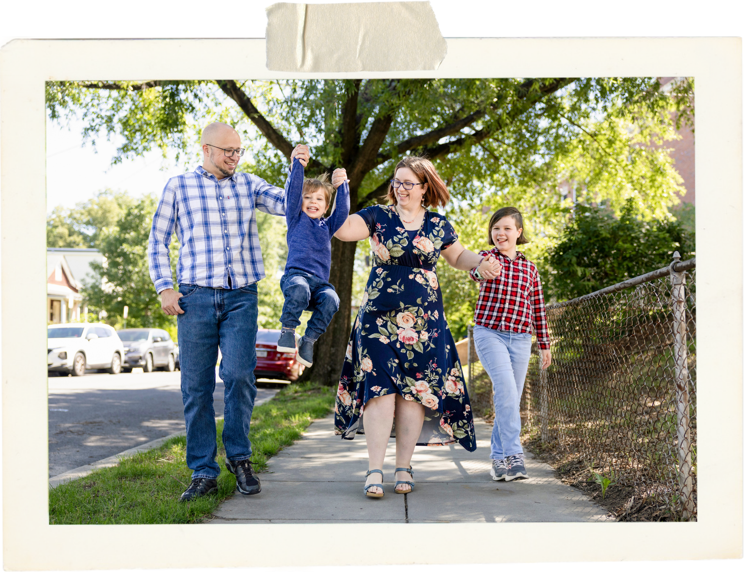 A family of four outdoors on a sidewalk, with green trees and parked cars in the background. The father and mother hold a child's hands, lifting him into the air, all smiling and happy.