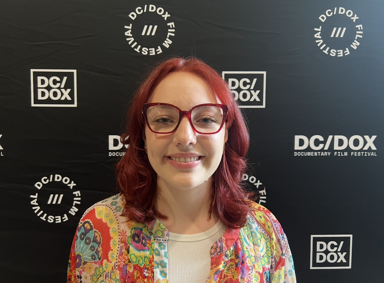 A smiling woman with red hair and large red glasses standing in front of a black backdrop with white logos for the DC/DOX Documentary Film Festival.