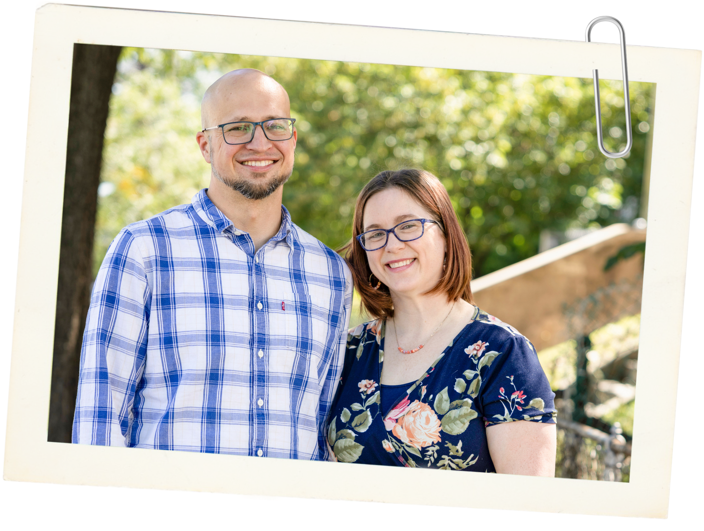 A man with glasses, a beard, and a bald head wearing a blue and white checkered shirt standing next to a woman with glasses, red hair, and a floral dress. They are outdoors with trees and a railing in the background.