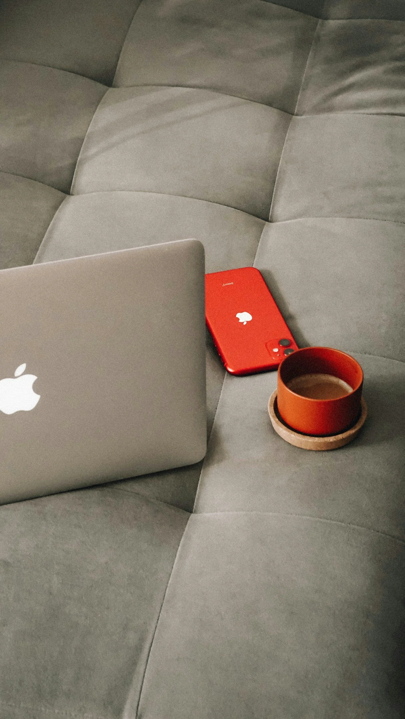 A gray couch with a silver MacBook, a red iPhone, and an orange cup of coffee on a tray.