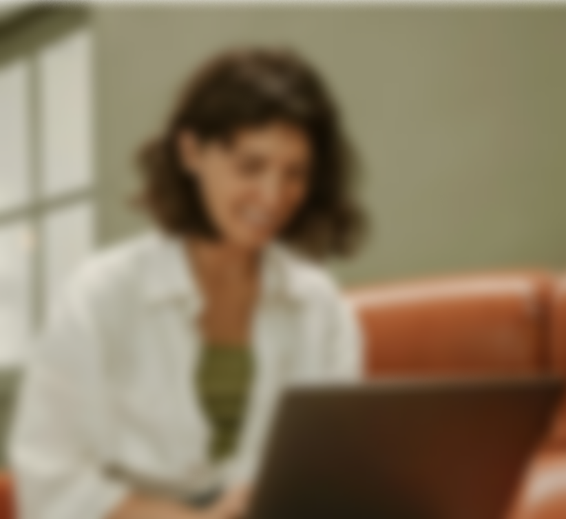 Blurry image of a woman with curly brown hair, wearing a white shirt, sitting in front of a laptop.