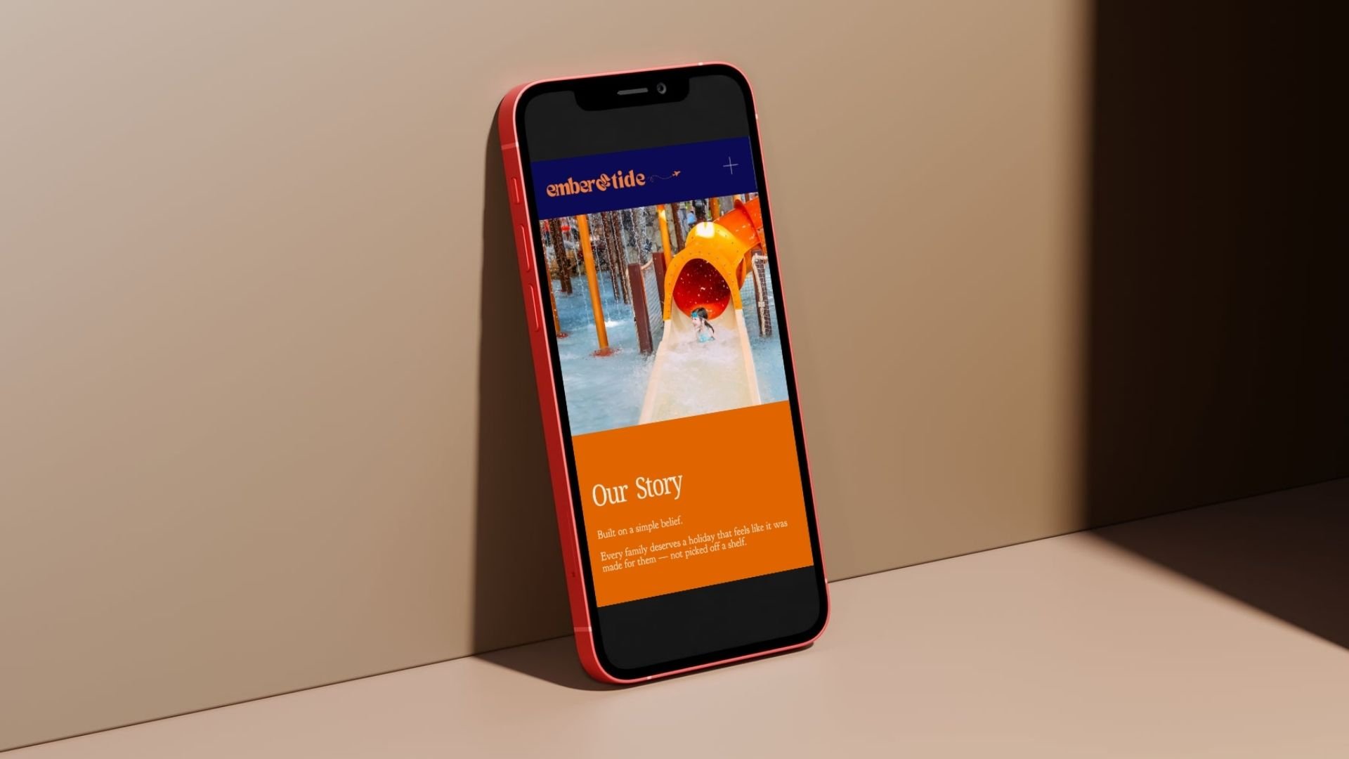 iPhone with a red frame leaning against a beige wall, displaying a webpage with a water slide, section titled 'Our Story', orange background, and text about building on a simple belief.