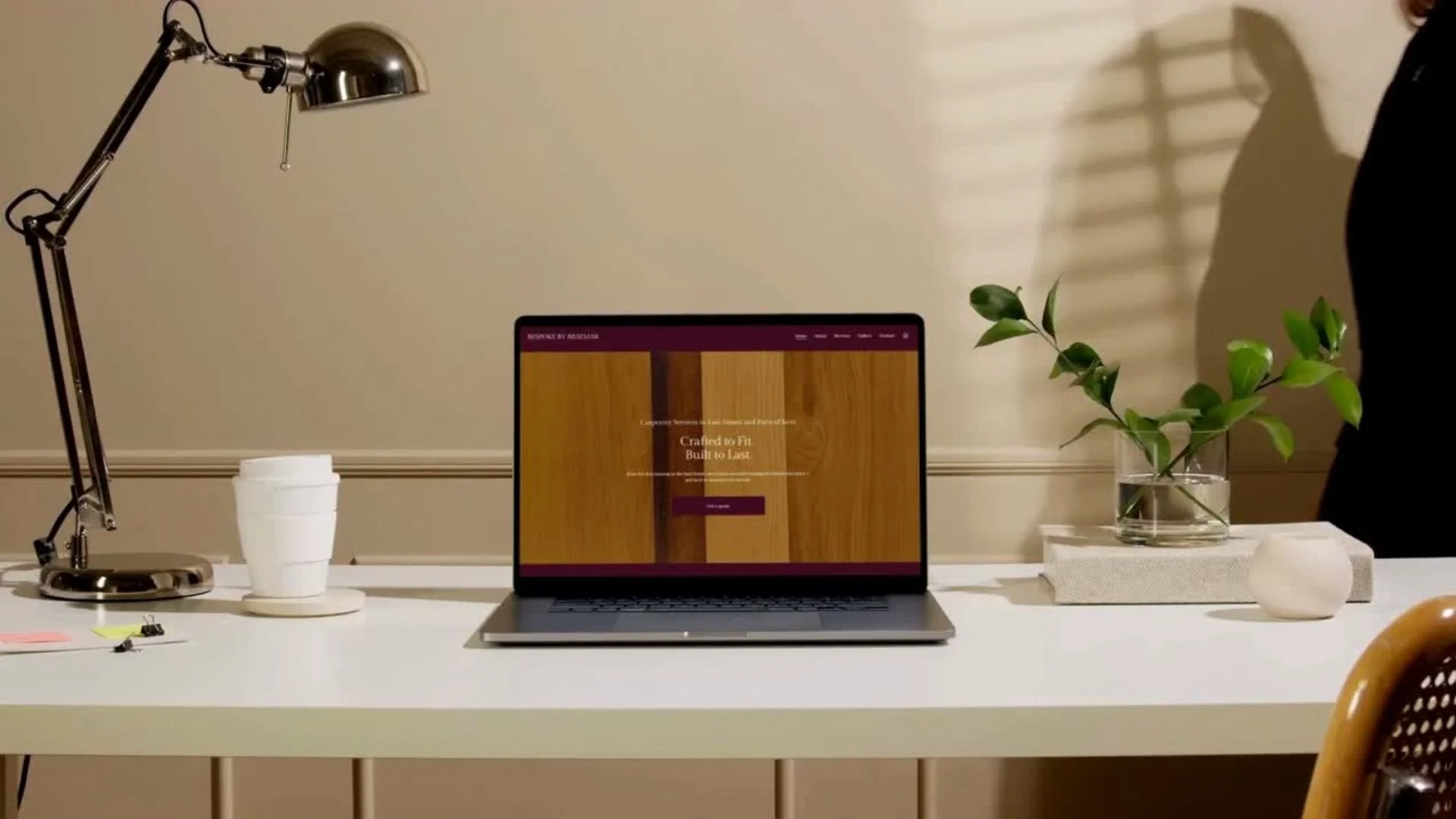 A white desk with a silver desk lamp, white ceramic cups, a laptop showing a website about wood products, a glass vase with a green plant, and a person partially visible on the right side of the image.