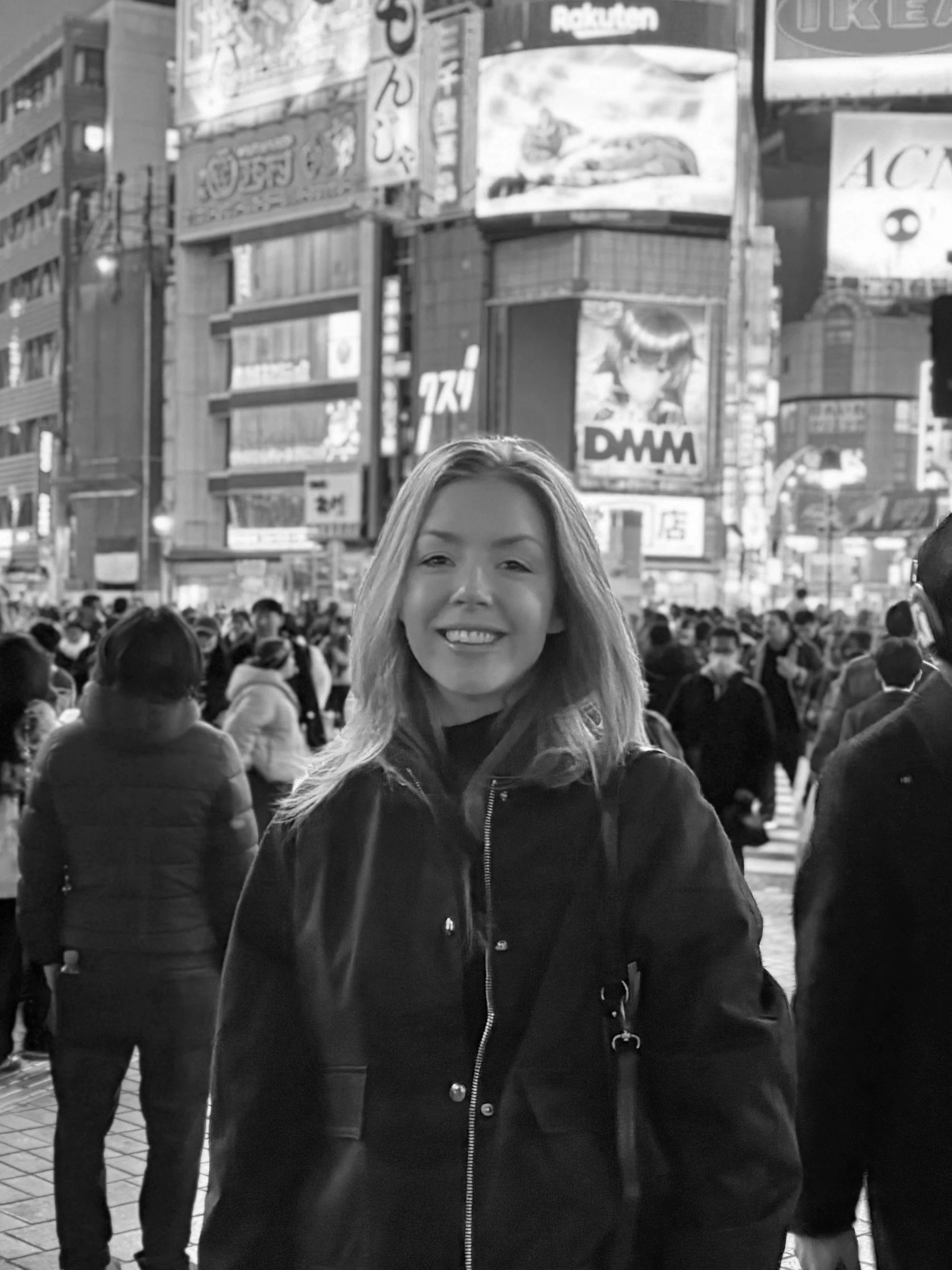 A smiling woman standing in a busy city square at night, with large illuminated billboards and advertisements in the background, and a crowd of people around her.