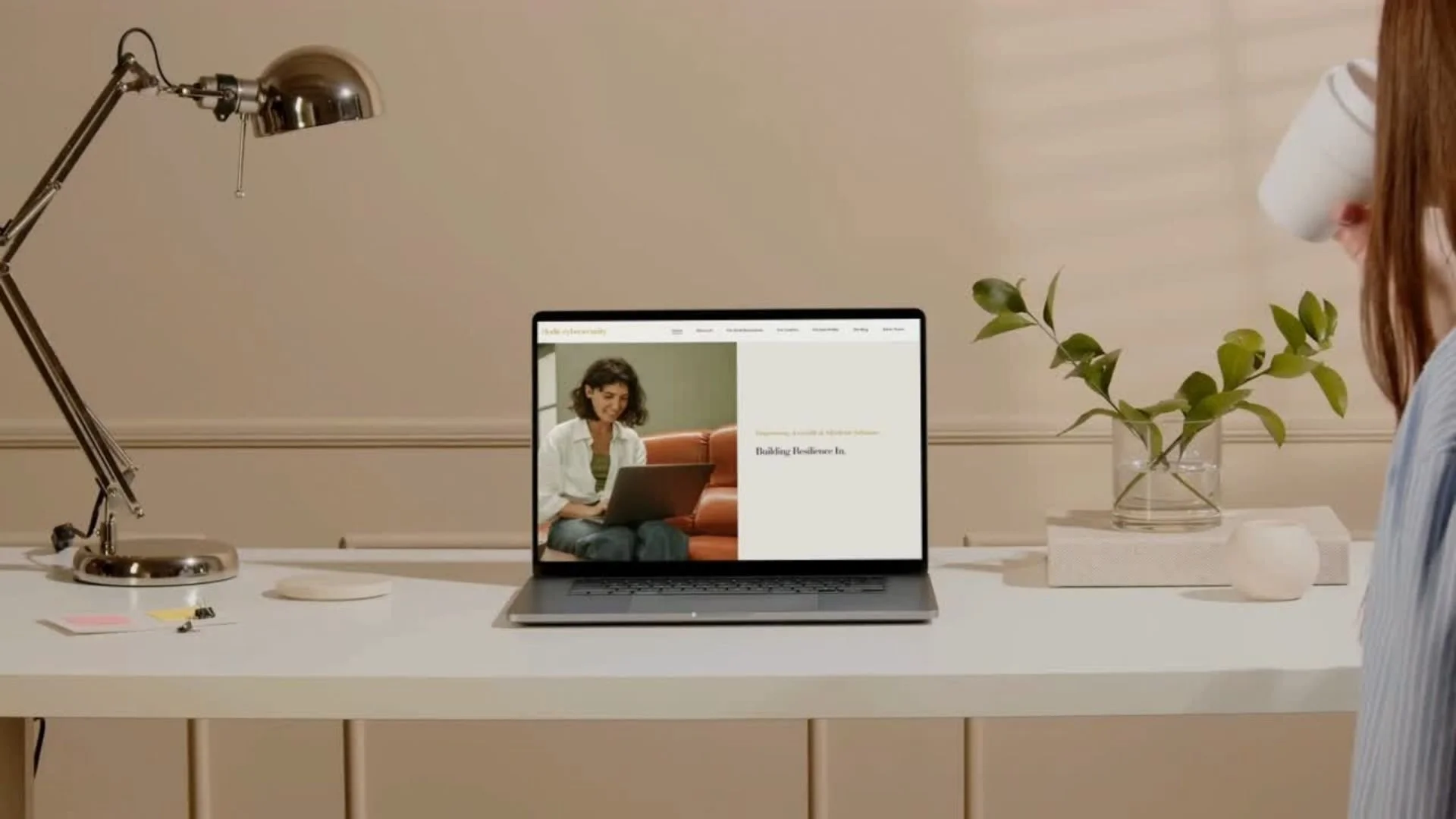 A modern workspace with a white desk, a silver adjustable desk lamp, a laptop displaying a woman sitting on a couch reading a book, a glass vase with green leafy plant, a white rectangular block, and a small round white container with a lid. A person
