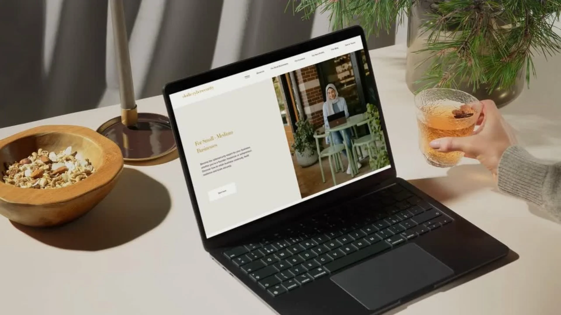 A person holding a glass of tea near a laptop displaying a website with a woman sitting at a table. On the table, there is a bowl of cereal, a lit candle, and a plant in a pot in the background.