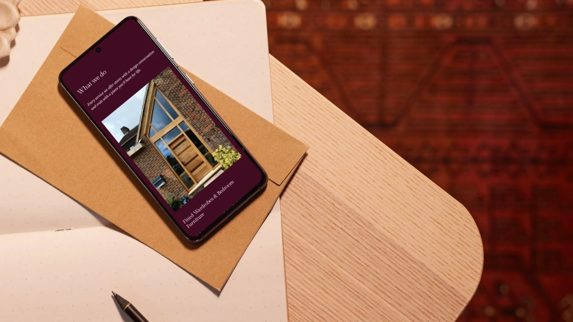 Smartphone on a table displaying a website about fitted wardrobes and bedroom furniture, with a brown envelope underneath and a black pen nearby. The table is light wood, with a white speckled surface nearby, and there is a patterned rug on the floor