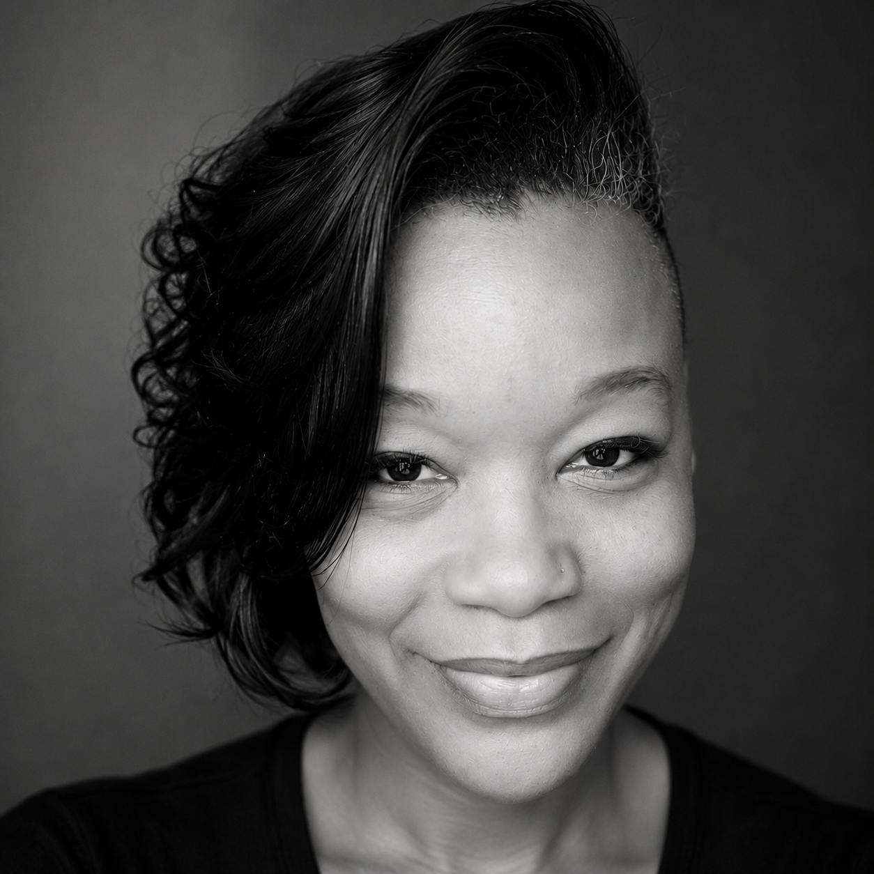 A black-and-white portrait of a woman with a short, wavy hairstyle, smiling softly at the camera, wearing a black top.
