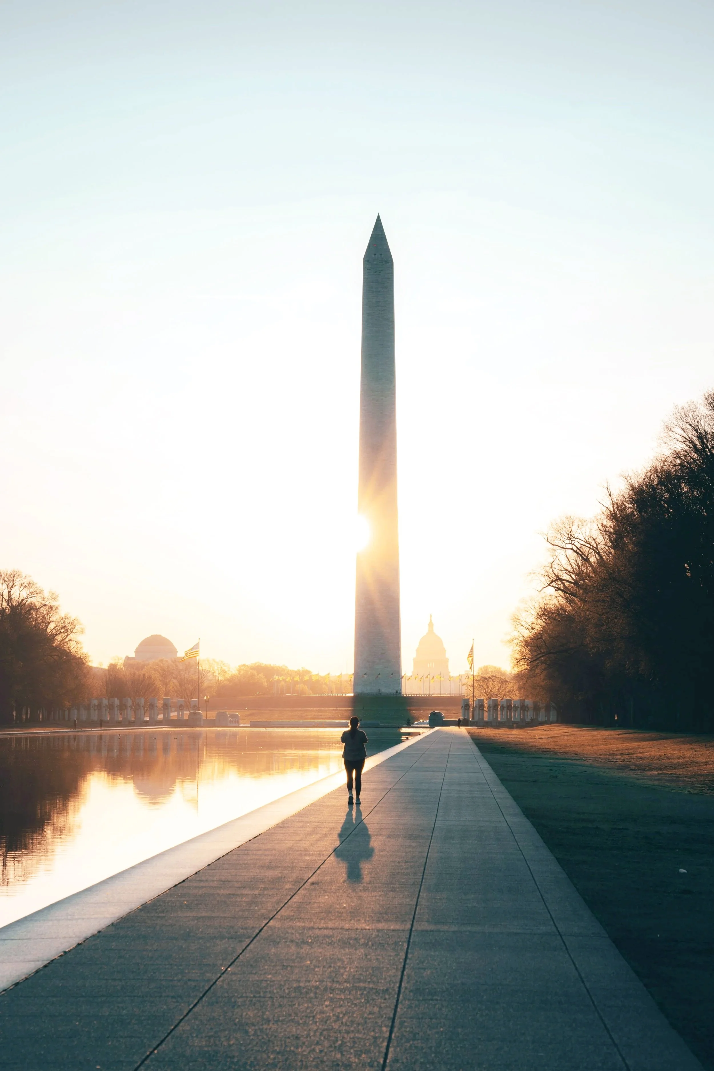 The Washington Monument is depicted in this photo taken during sunrise or sunset, with a person walking along the reflecting pool in front of it. The scene features a clear sky and some trees on the sides.