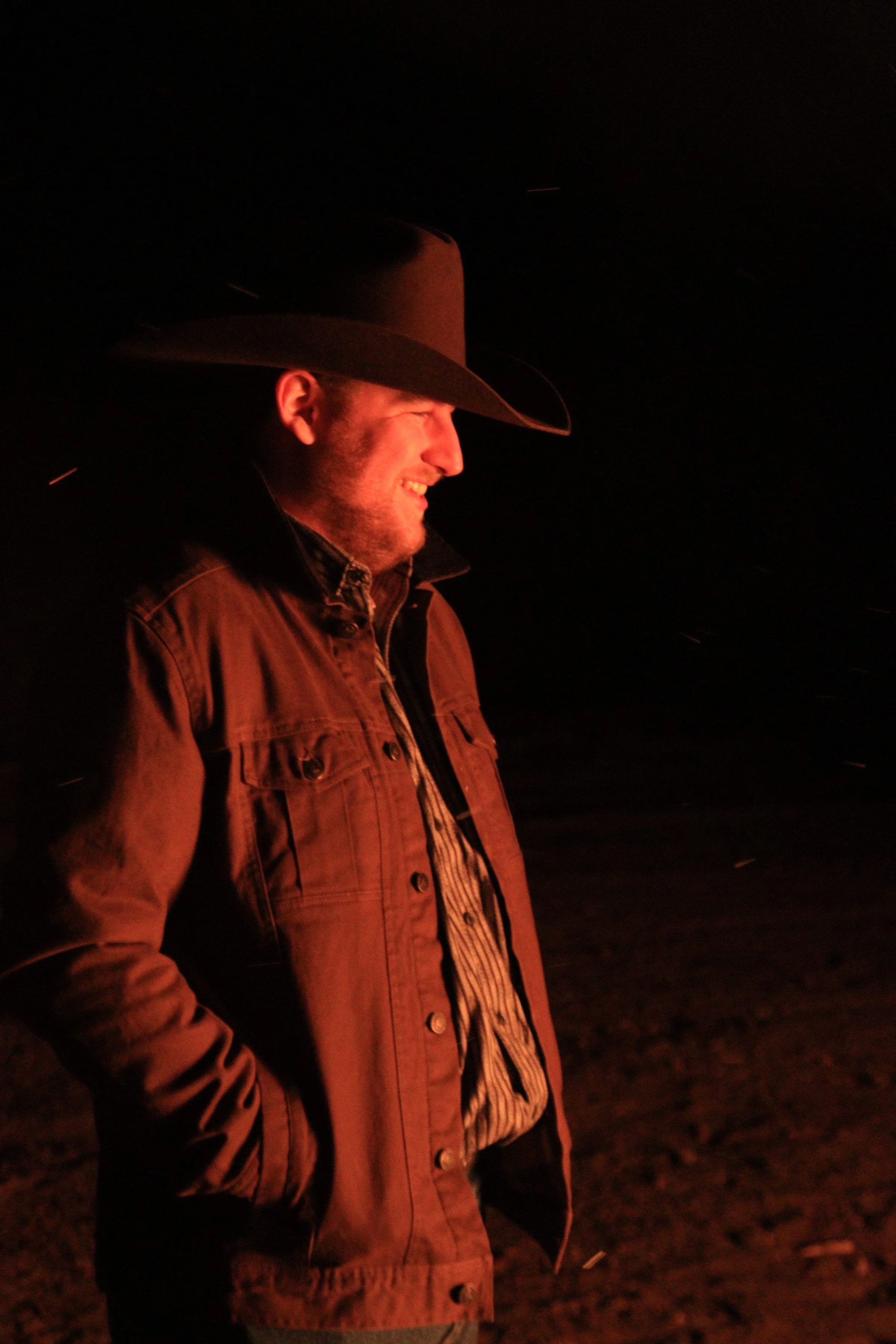 A man is smiling and wearing a wide-brimmed hat and a brown jacket, standing outdoors at night illuminated by a warm light.