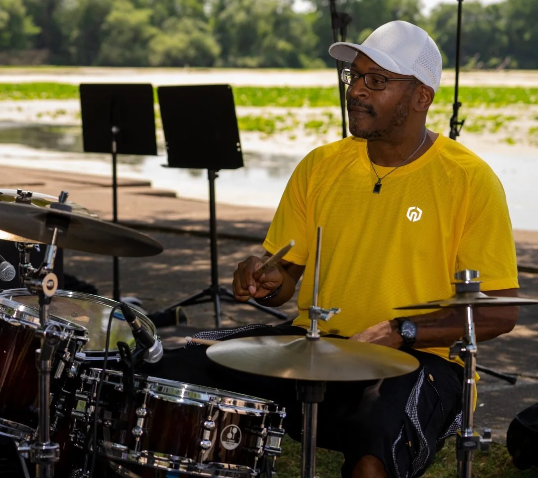 A man in a yellow shirt, glasses, and a white cap playing drums outdoors near a lake with greenery in the background.