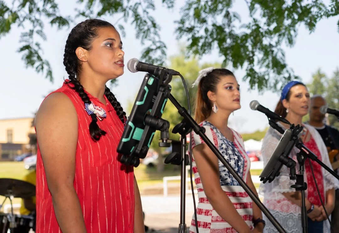 Group of four women singing into microphones outdoors, with trees and buildings in the background.