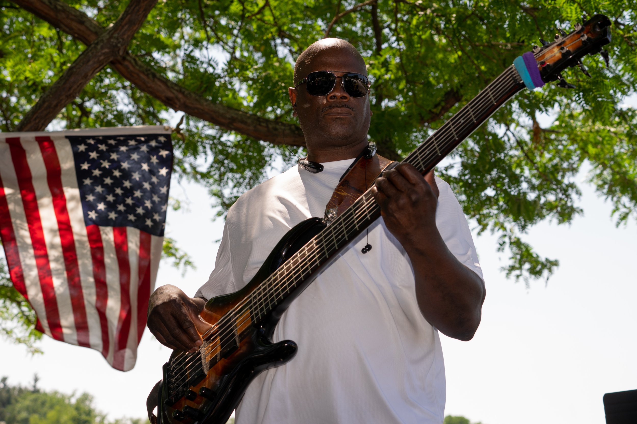 An African American man wearing sunglasses and a white shirt playing an electric guitar outdoors under a leafy tree, with an American flag hanging nearby.