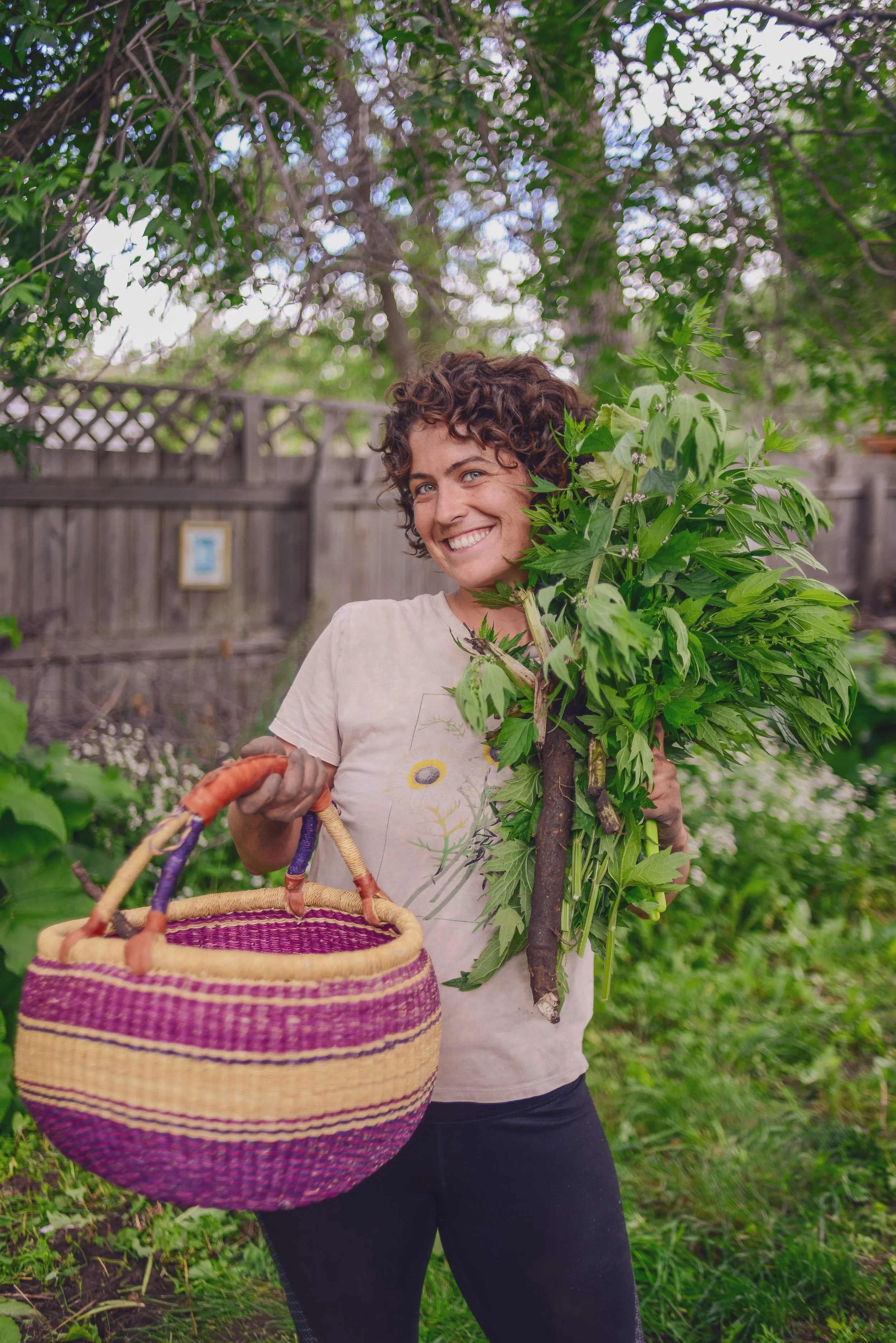 A woman with short, curly dark hair smiling outdoors, holding a large leafy plant and a colorful woven basket, with a wooden fence and trees in the background.